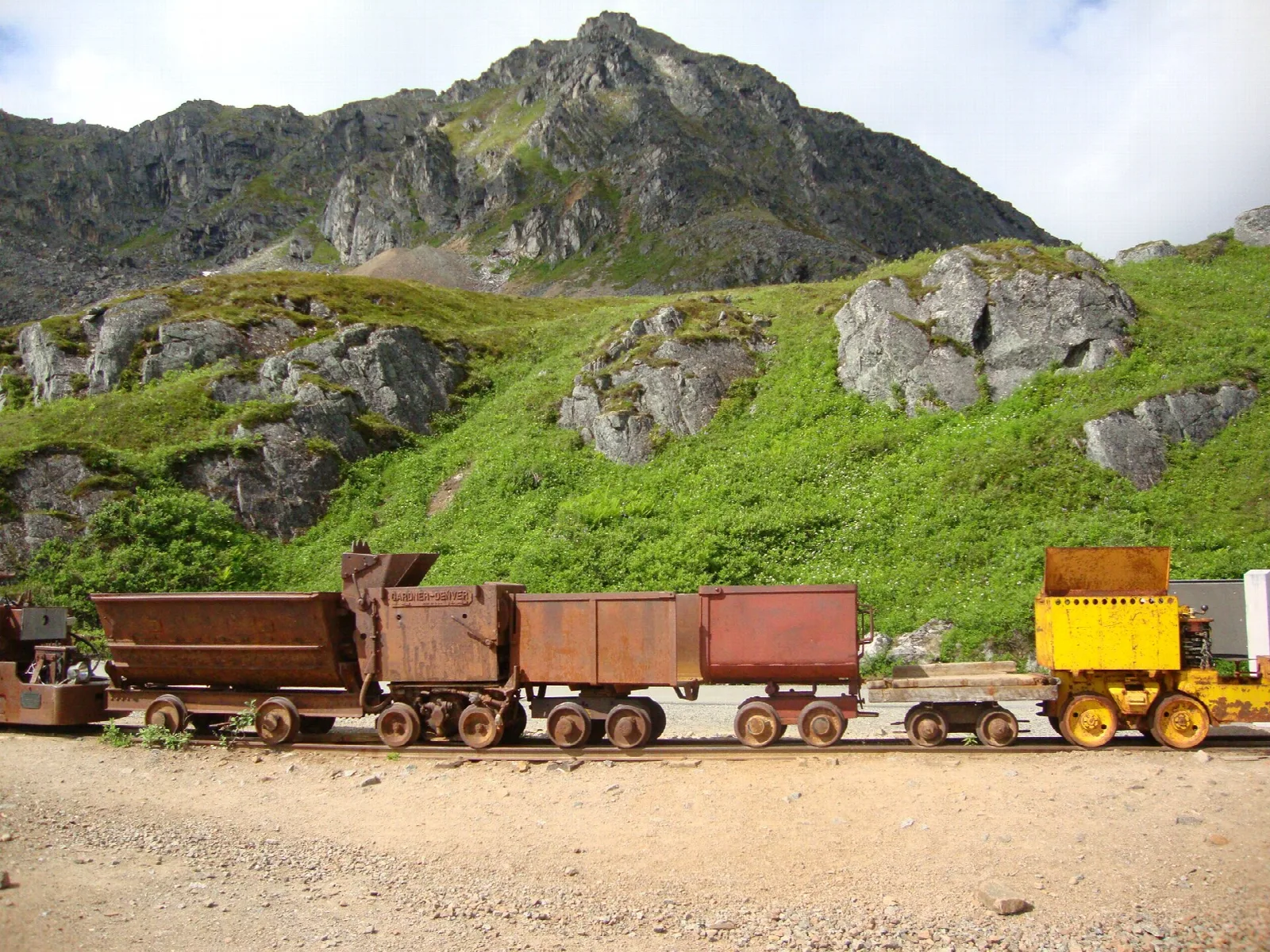 Row of rusty ore cars on a gravel track below a rocky green slope