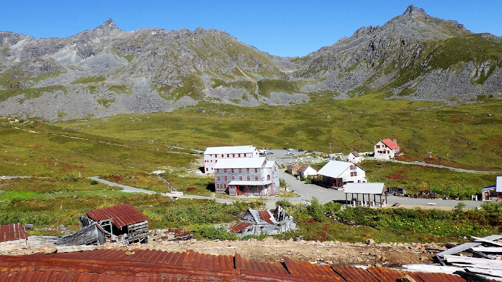 Old white mine buildings scattered across a green mountain valley under jagged peaks