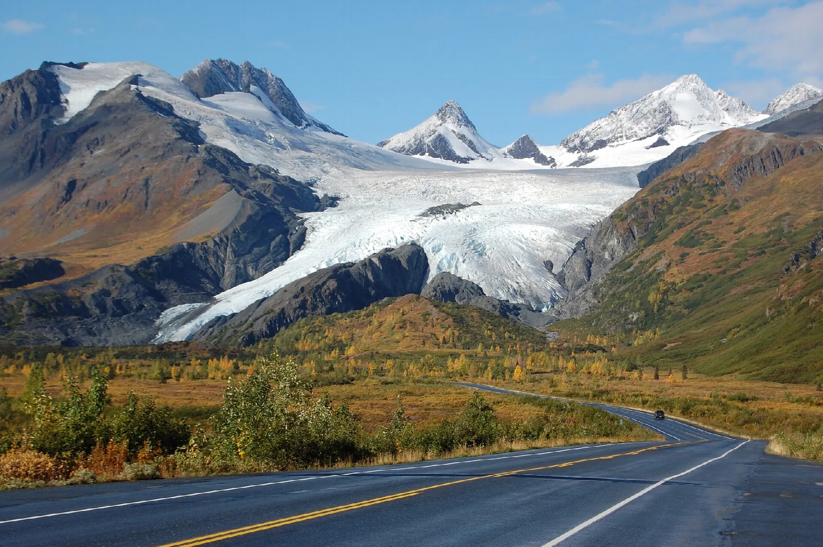 Glacier between snowy peaks above a highway curving through autumn tundra