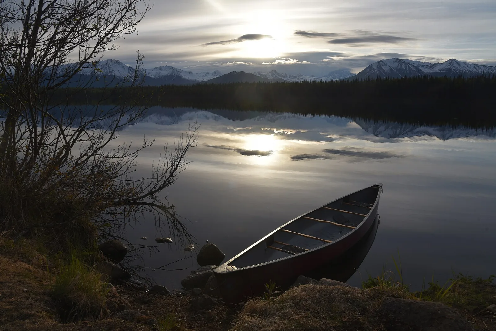 A canoe on the shore of a still lake at sunset with mountains in the distance