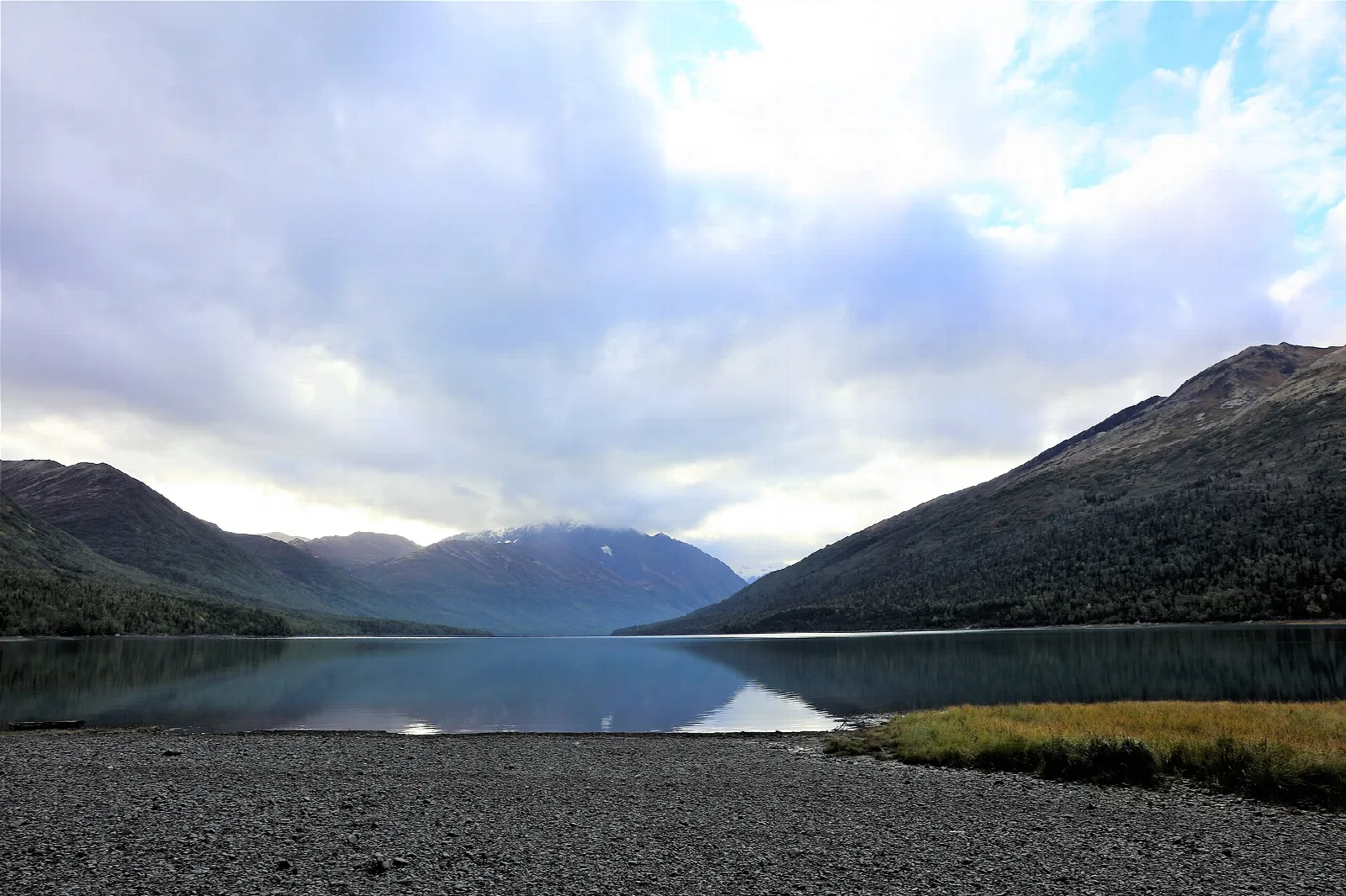 A gravel shore along a still lake with mountains in the distance