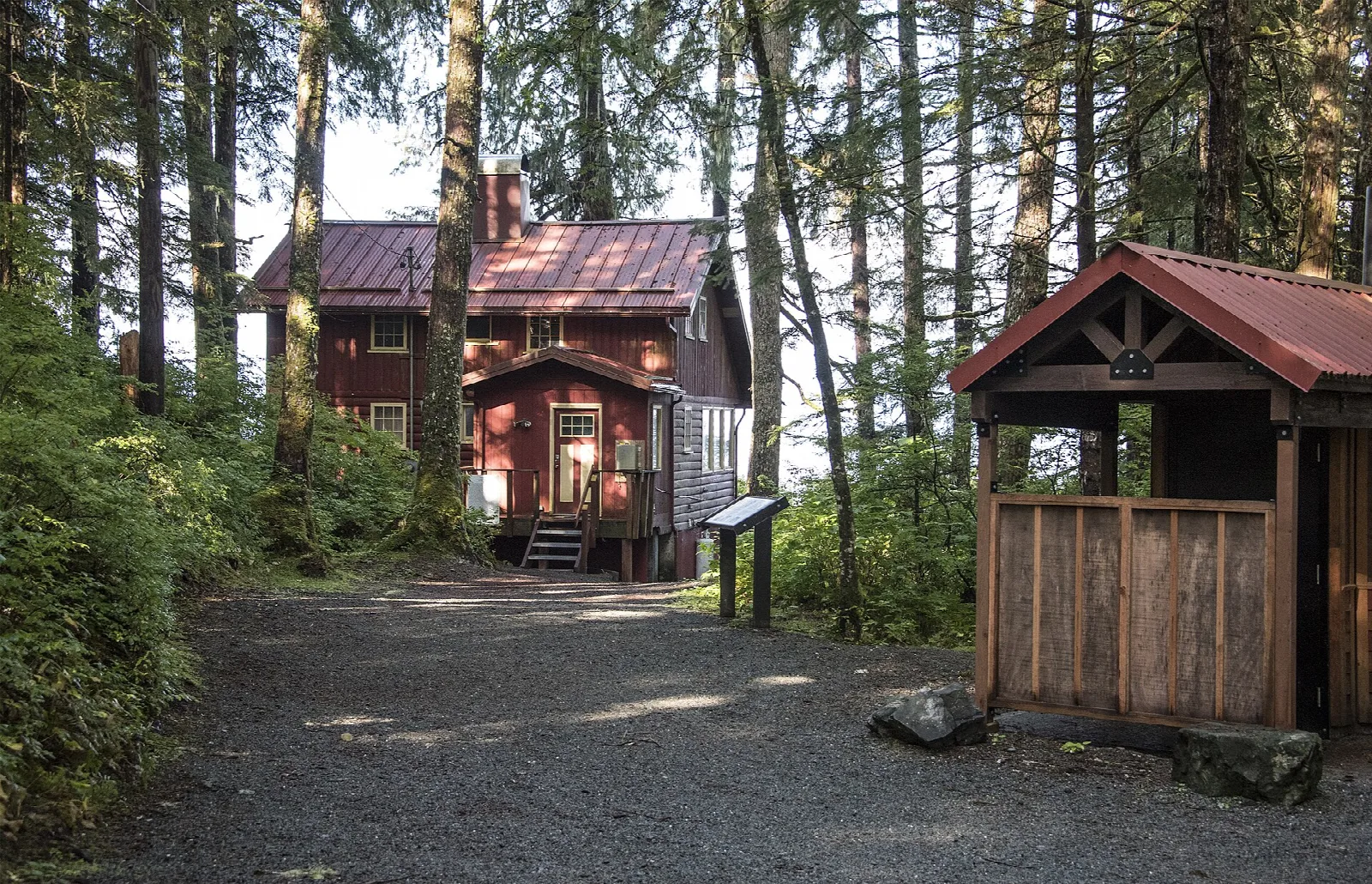 A red log cabin and small shelter among tall spruce along a gravel path