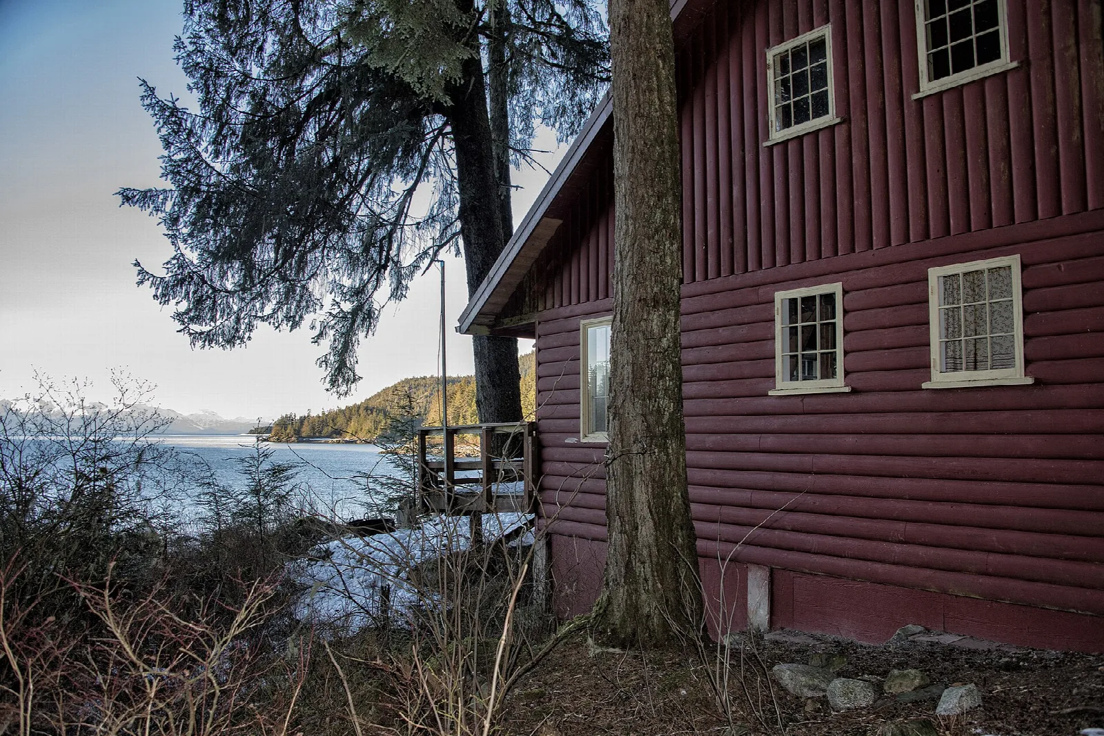 Side of a red cabin among trees with water visible through the forest