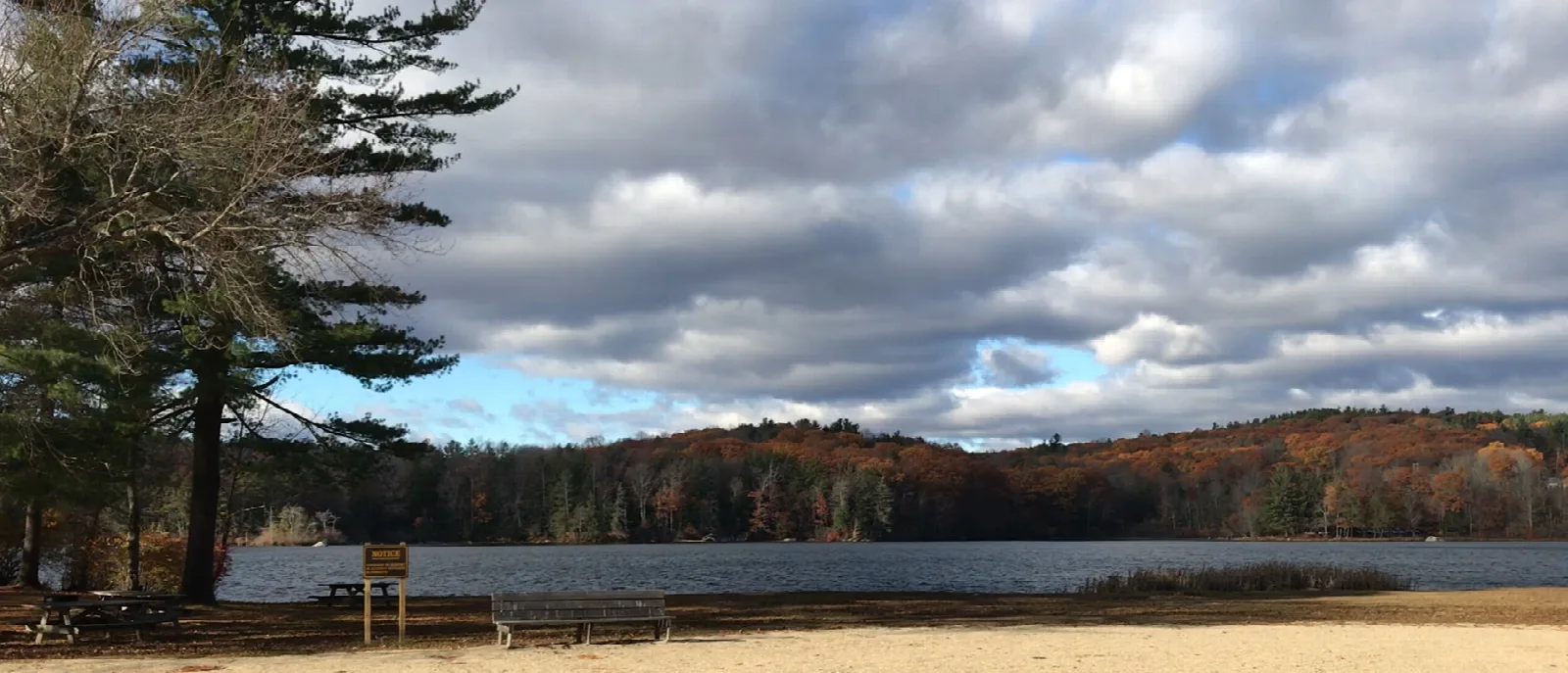 A sandy shore and a pond below forested hills under a cloudy sky