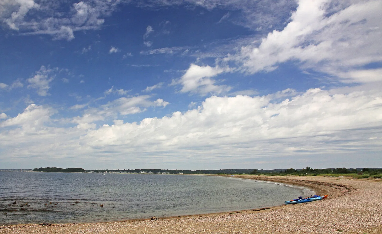 A wide gravel beach curving along calm water under a cloudy sky