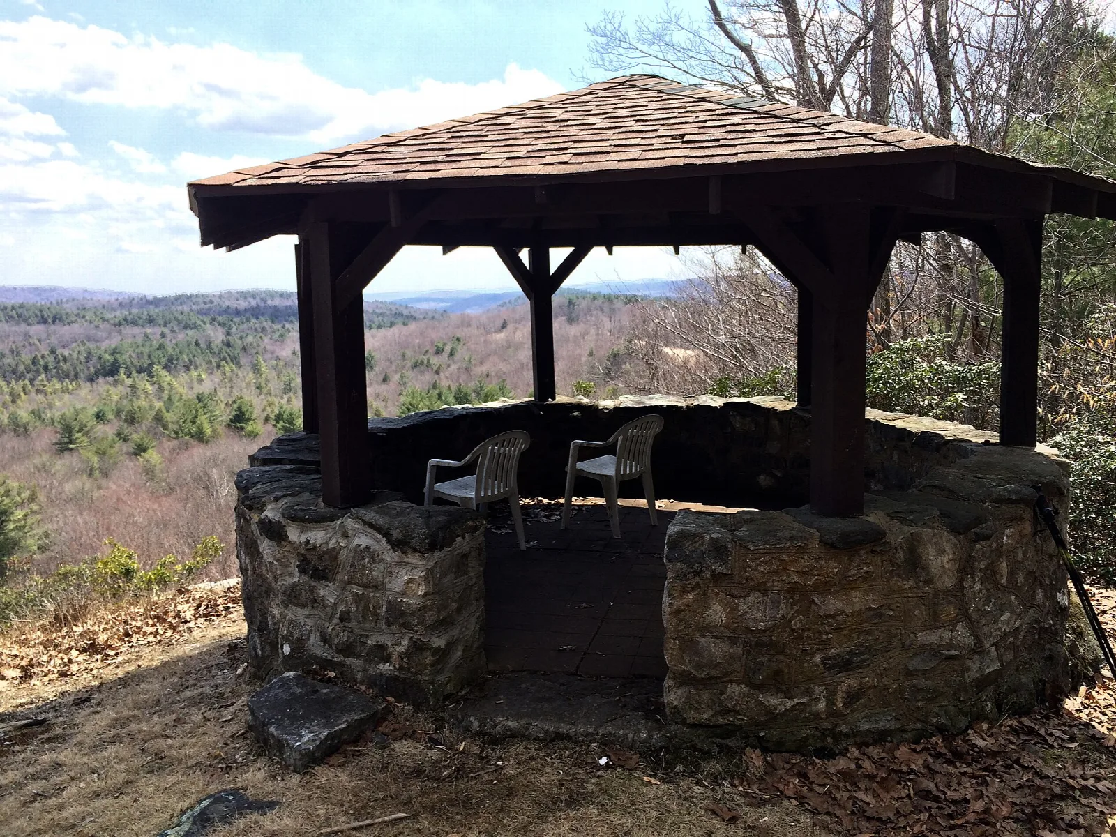 Open stone-walled shelter with a shingle roof and chairs, hills and forest beyond