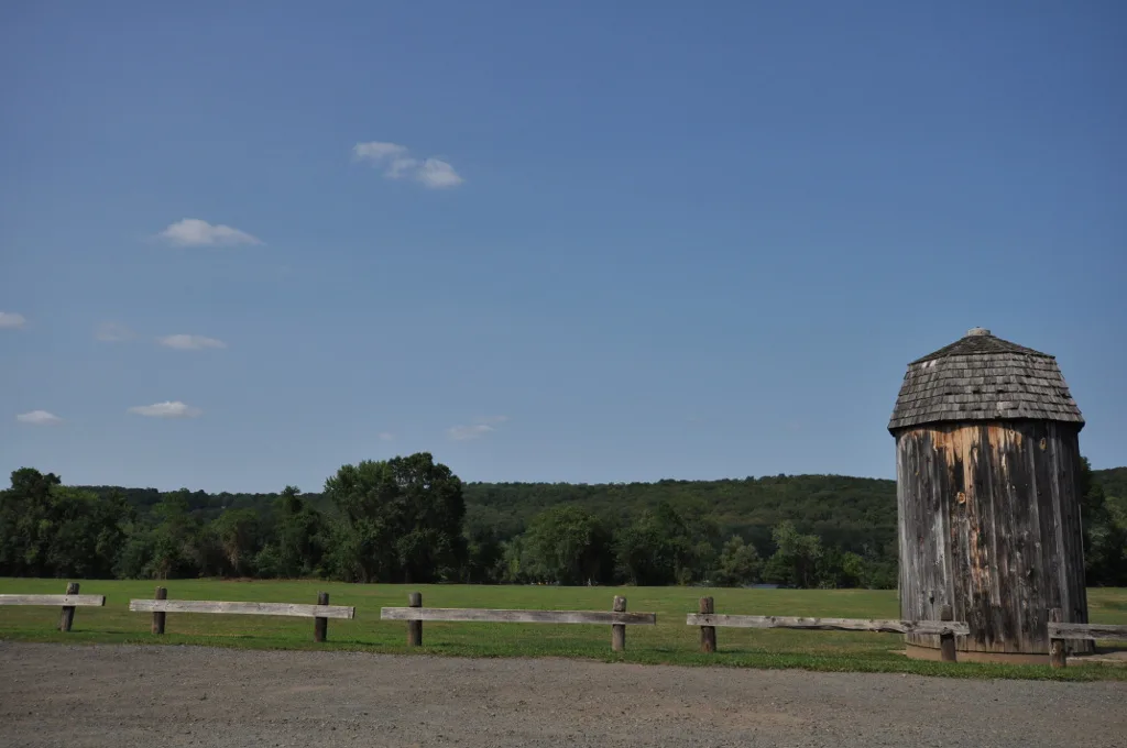 Weathered wooden tower with a shingled cap beside a grass field and rail fence under blue sky