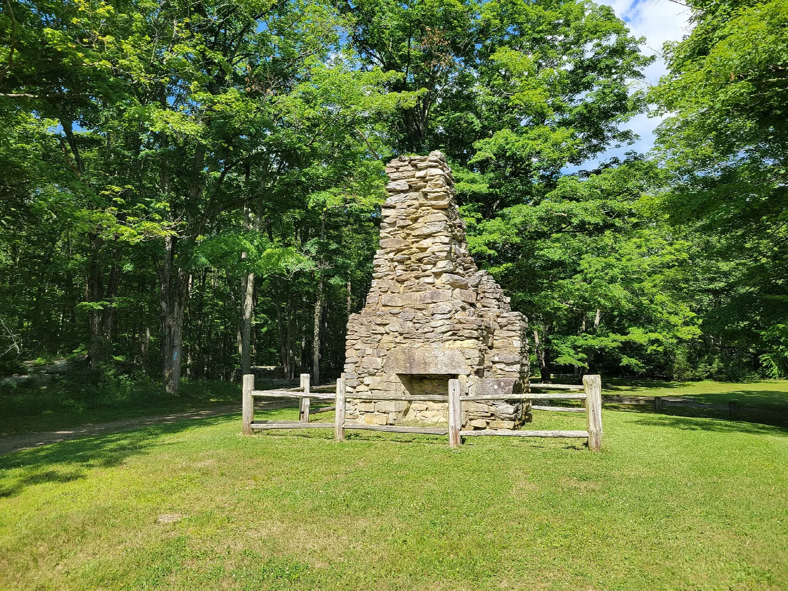 Tall stone chimney on a lawn behind a wooden fence, surrounded by trees