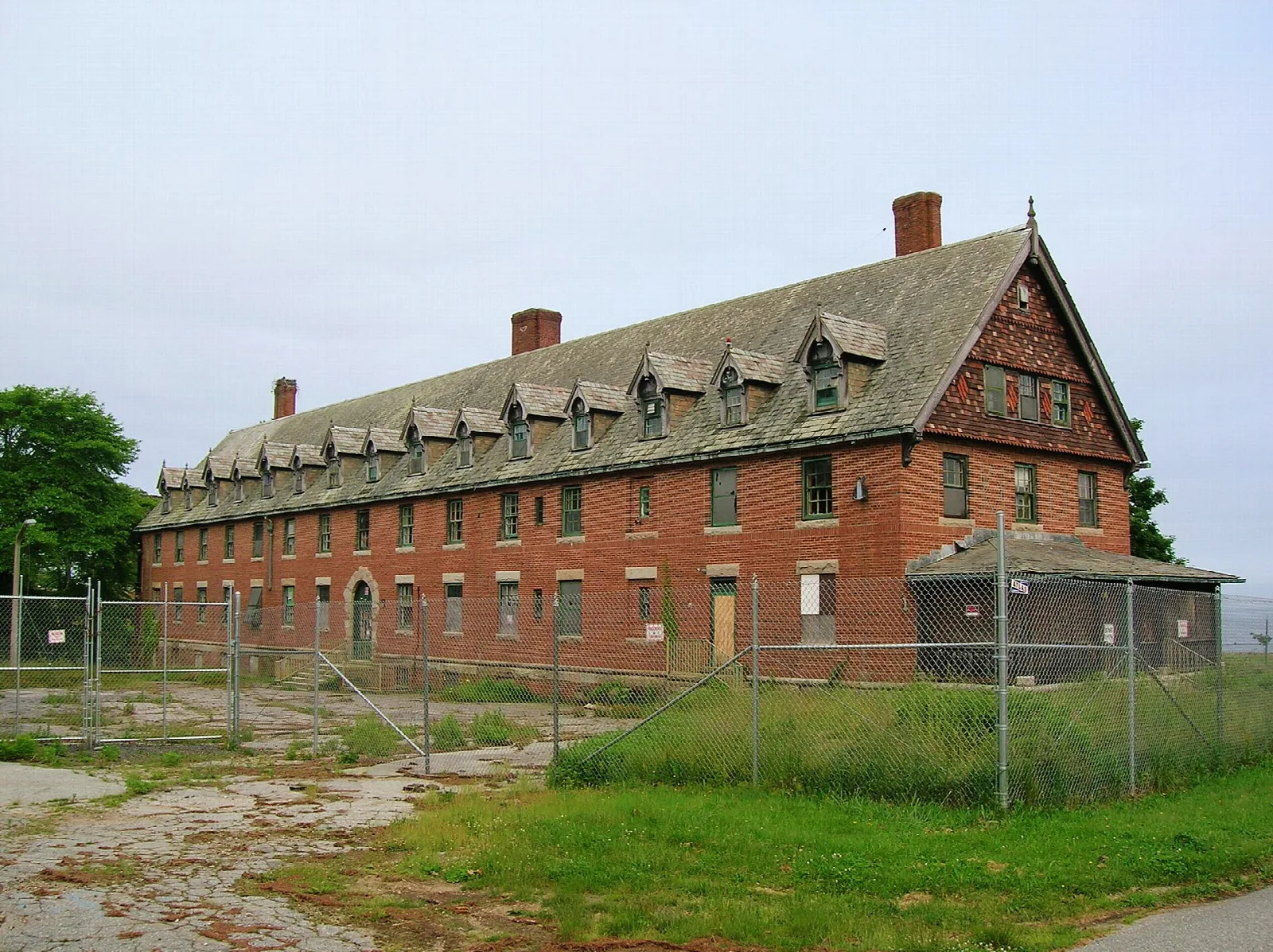 Long brick building with a steep dormered roof behind a fence