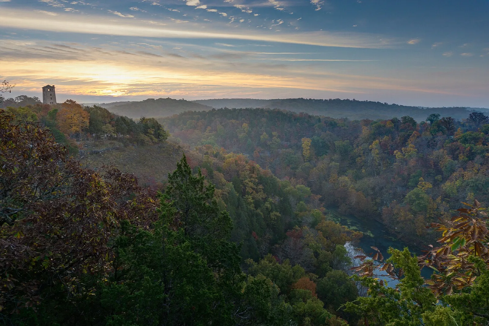 Forested river valley seen from a hilltop at sunset, with a distant stone tower on the ridge
