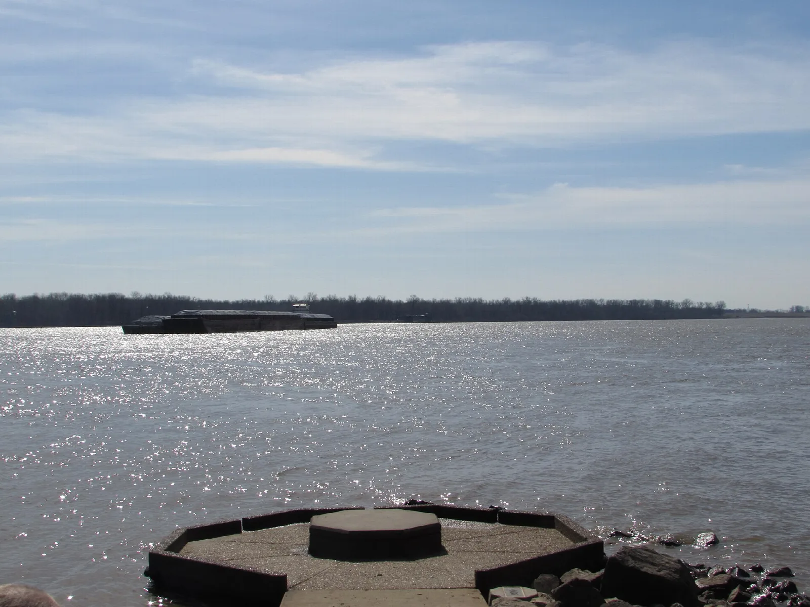 Round stone platform at the river edge with a barge on the far horizon