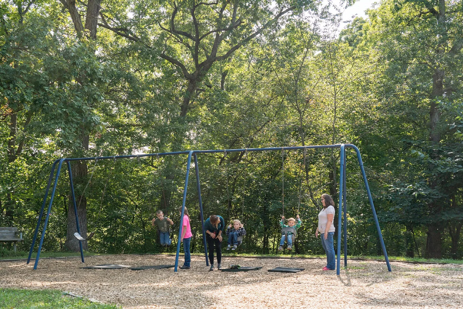 Children and adults at a swing set on a gravel play area under trees