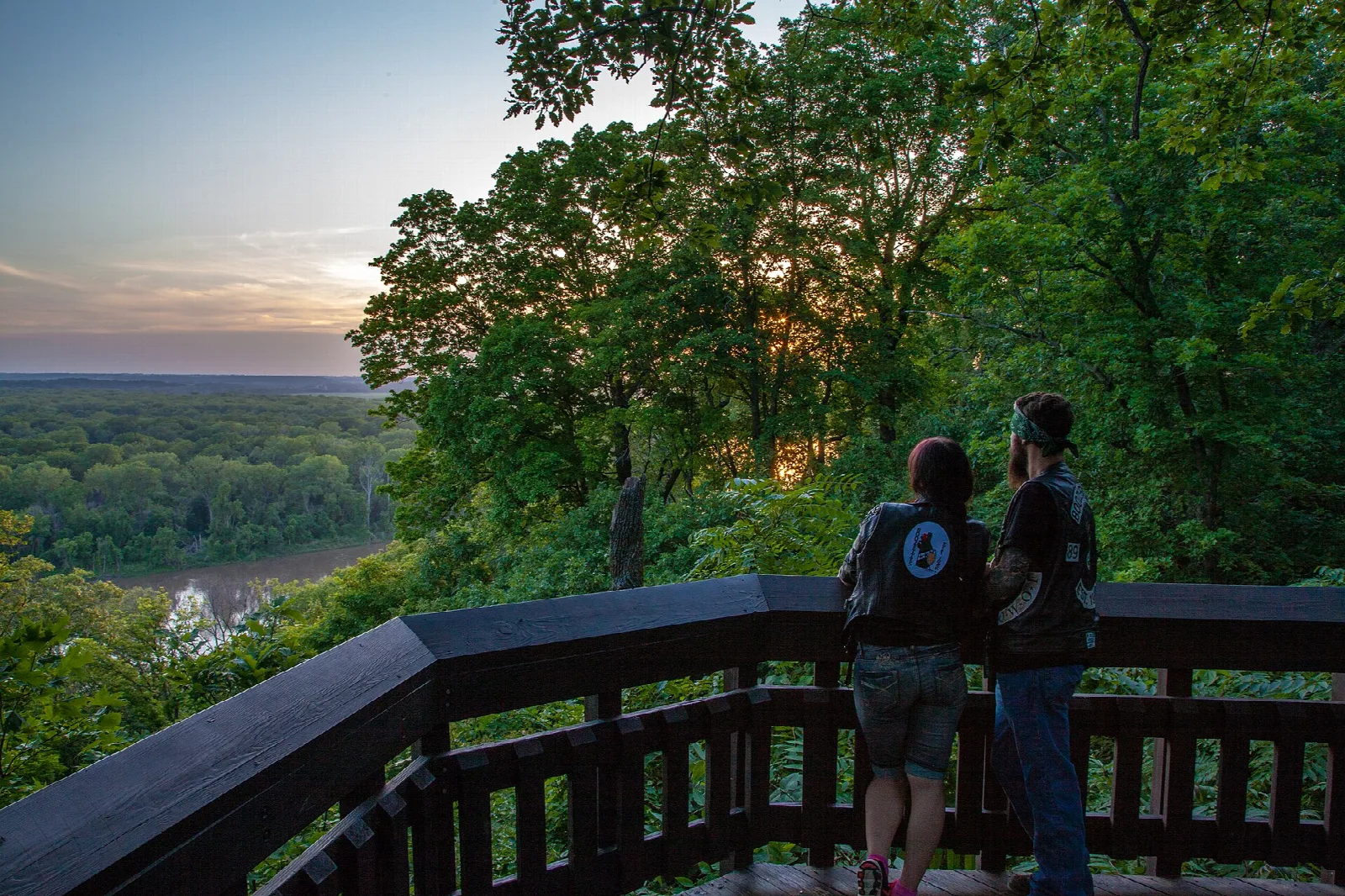 Two people at a wooden railing looking over a wooded river valley at sunset