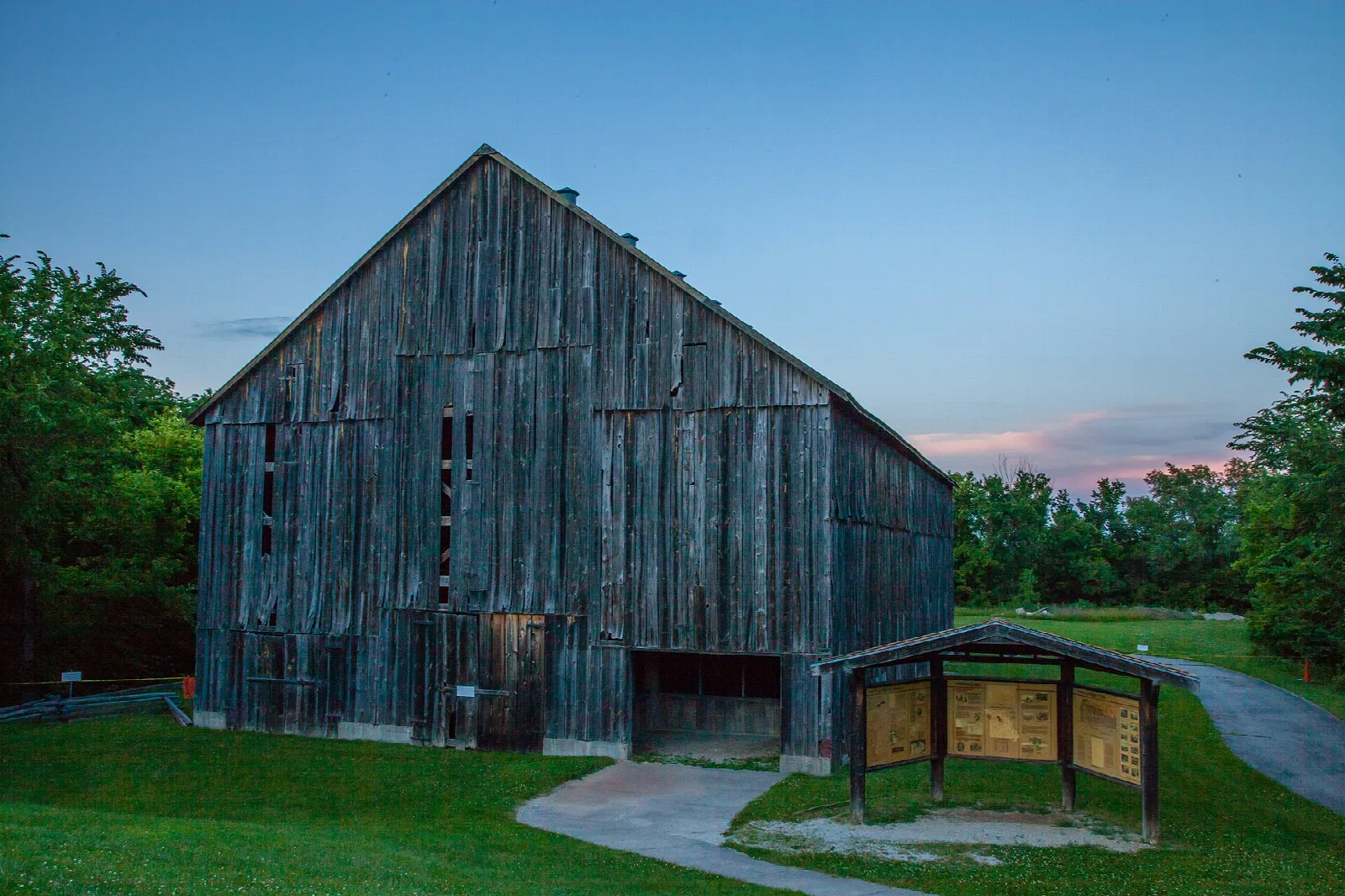 Weathered wooden barn with an information kiosk and a paved path at dusk