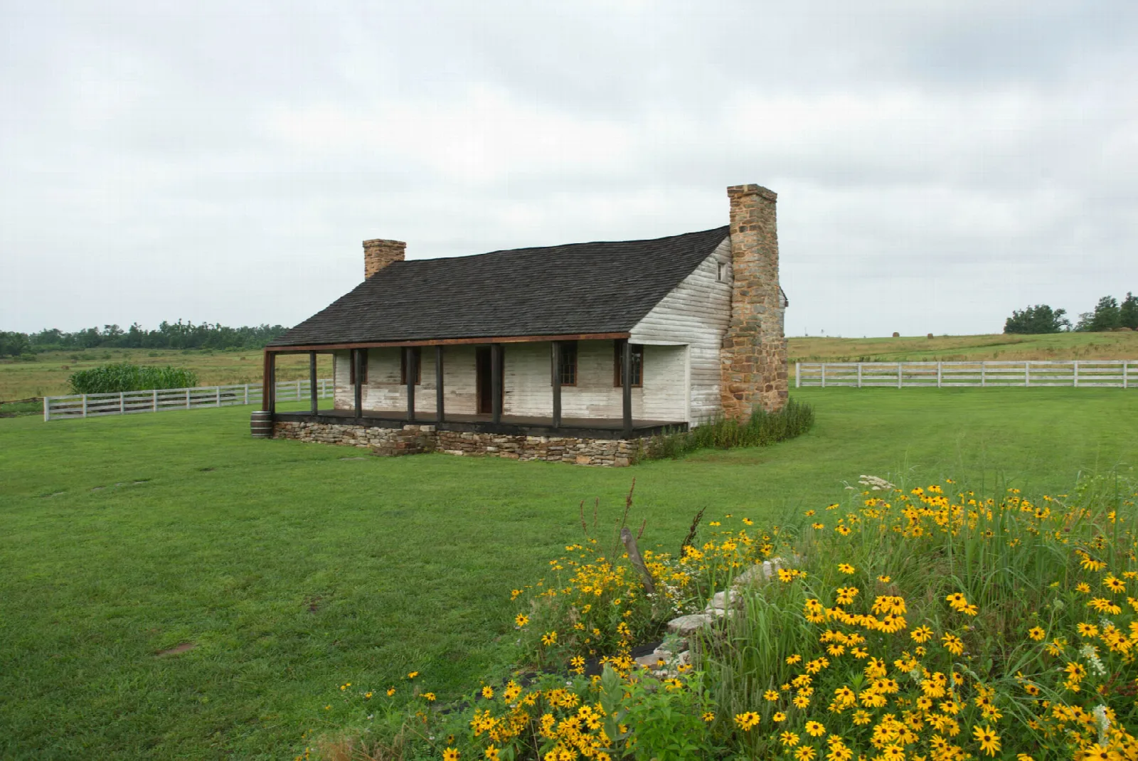 Low stone-chimney house with a long porch on a green lawn, yellow wildflowers in front