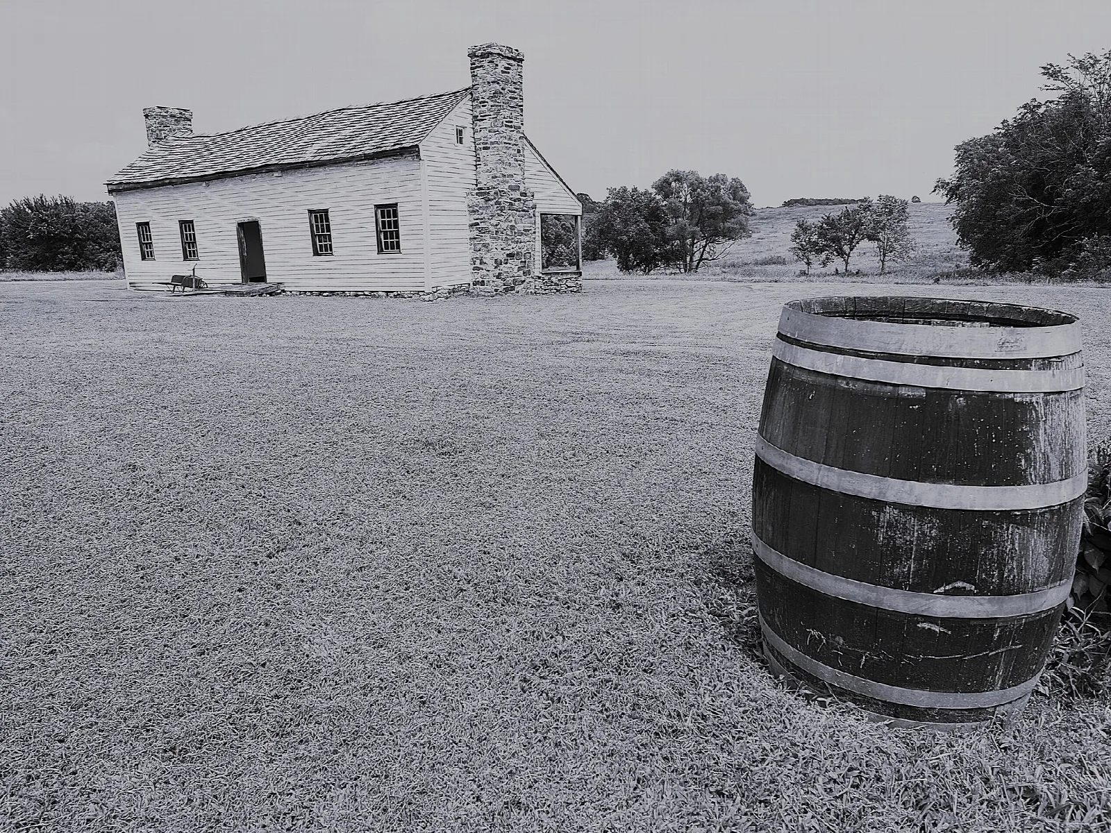 Black and white view of a stone-chimney house across a lawn with a wooden barrel in front