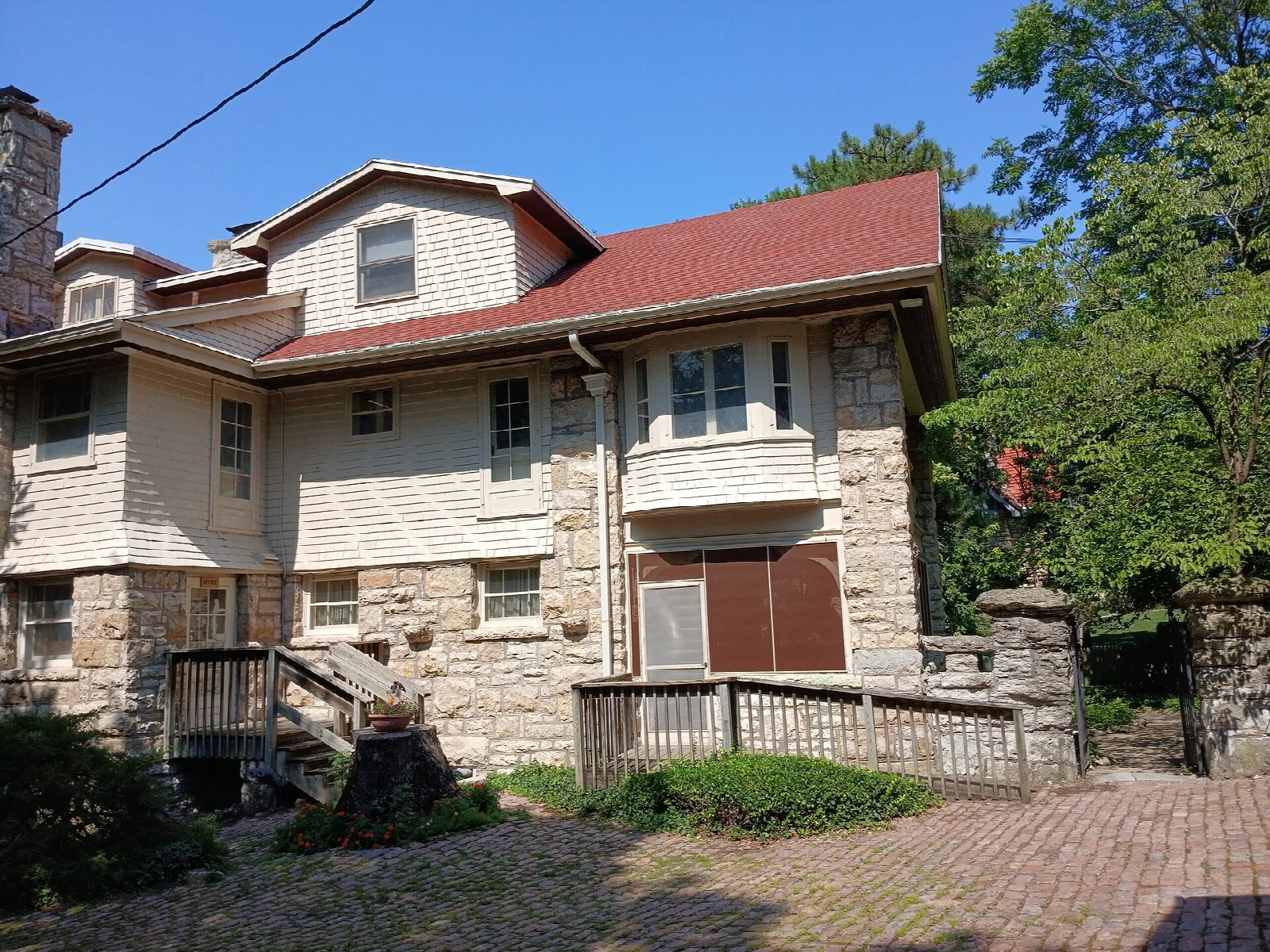 Stone and wood house with a red roof seen from a brick drive, trees around it