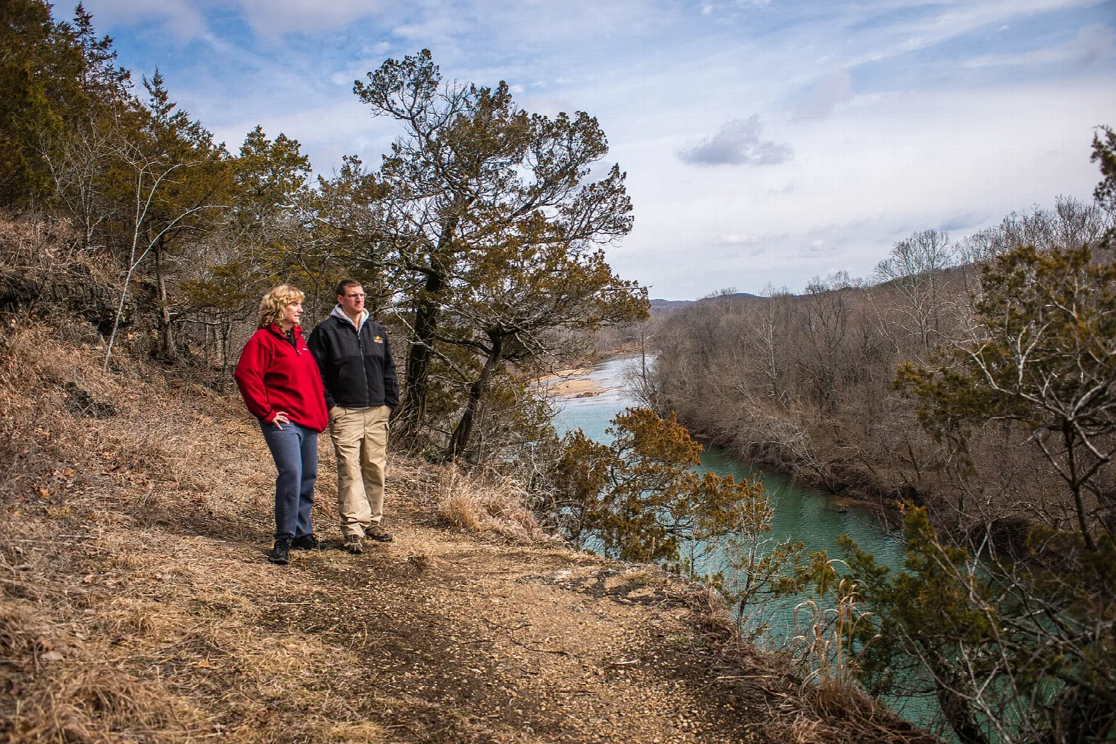 Two people on a dry hillside looking out over a turquoise river and wooded bluffs