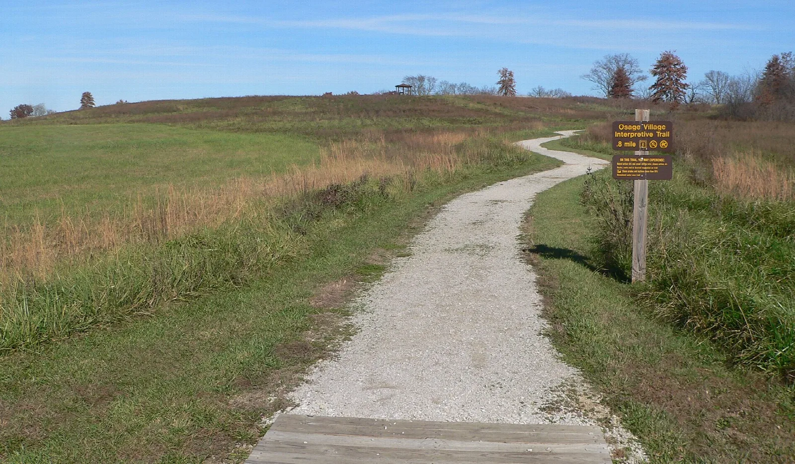 Gravel path climbing a grassy hill toward a trail sign under blue sky