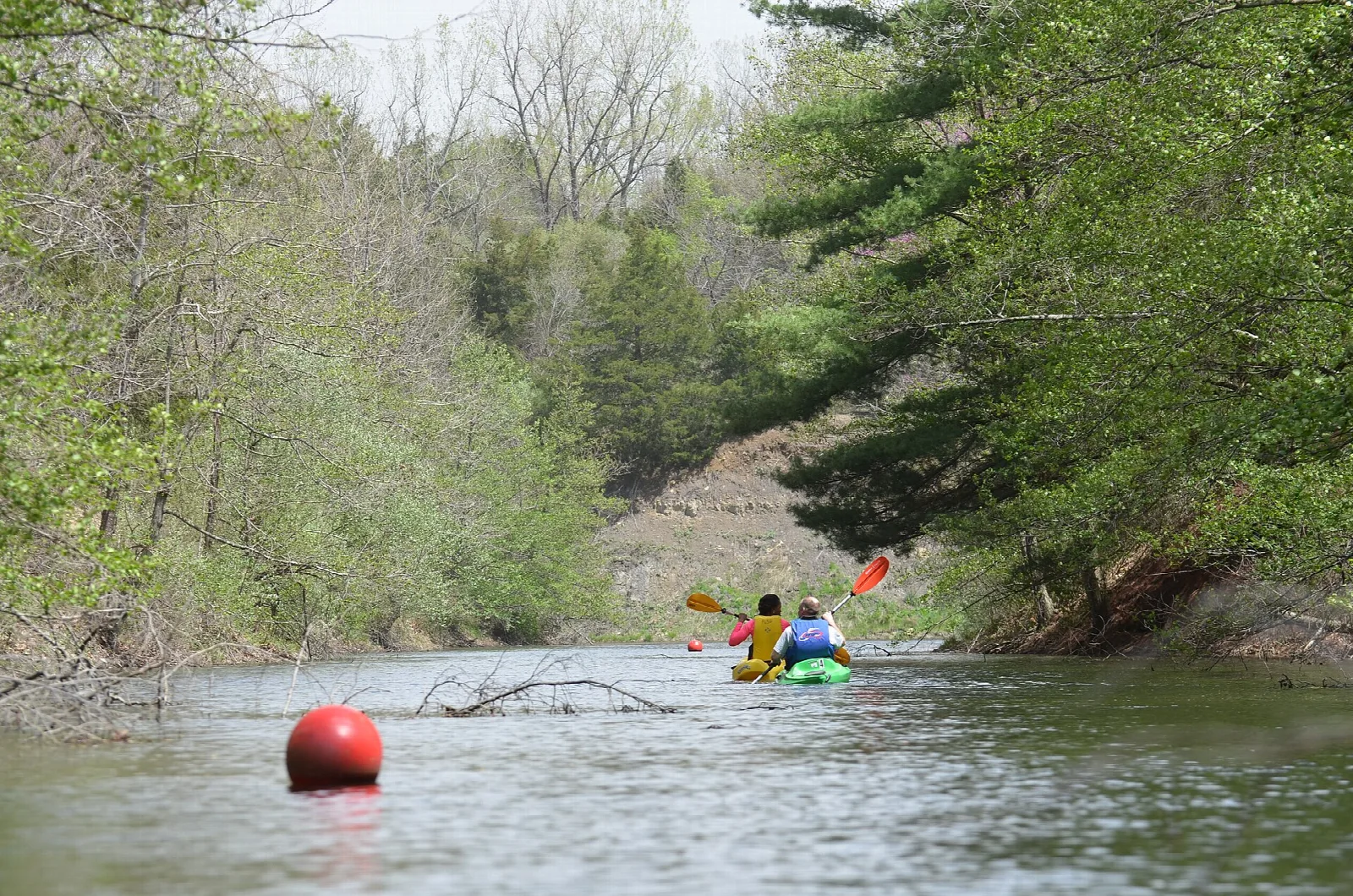 Two people paddling kayaks on a quiet lake past a red buoy and wooded banks