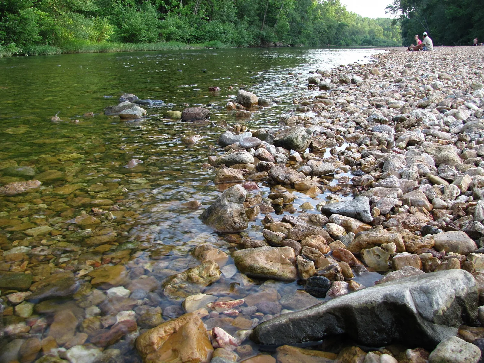 Clear shallow river over a bar of rounded rocks, with trees along the far bank