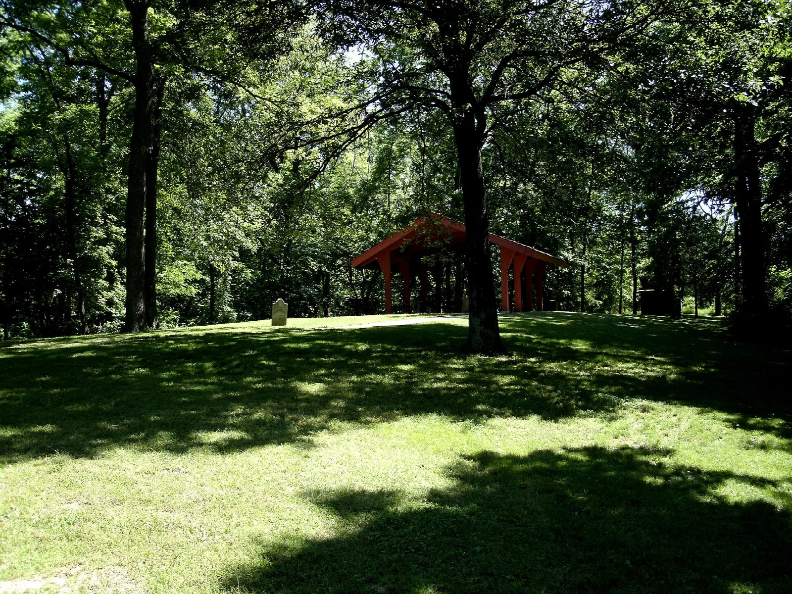 Mowed lawn under tall trees with a small red-roofed shelter in the distance