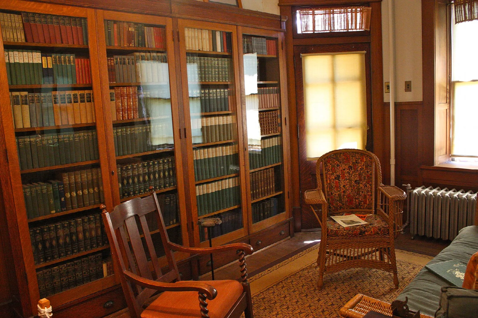 Library room with tall glass-front bookcases, wooden chairs and a sunlit window