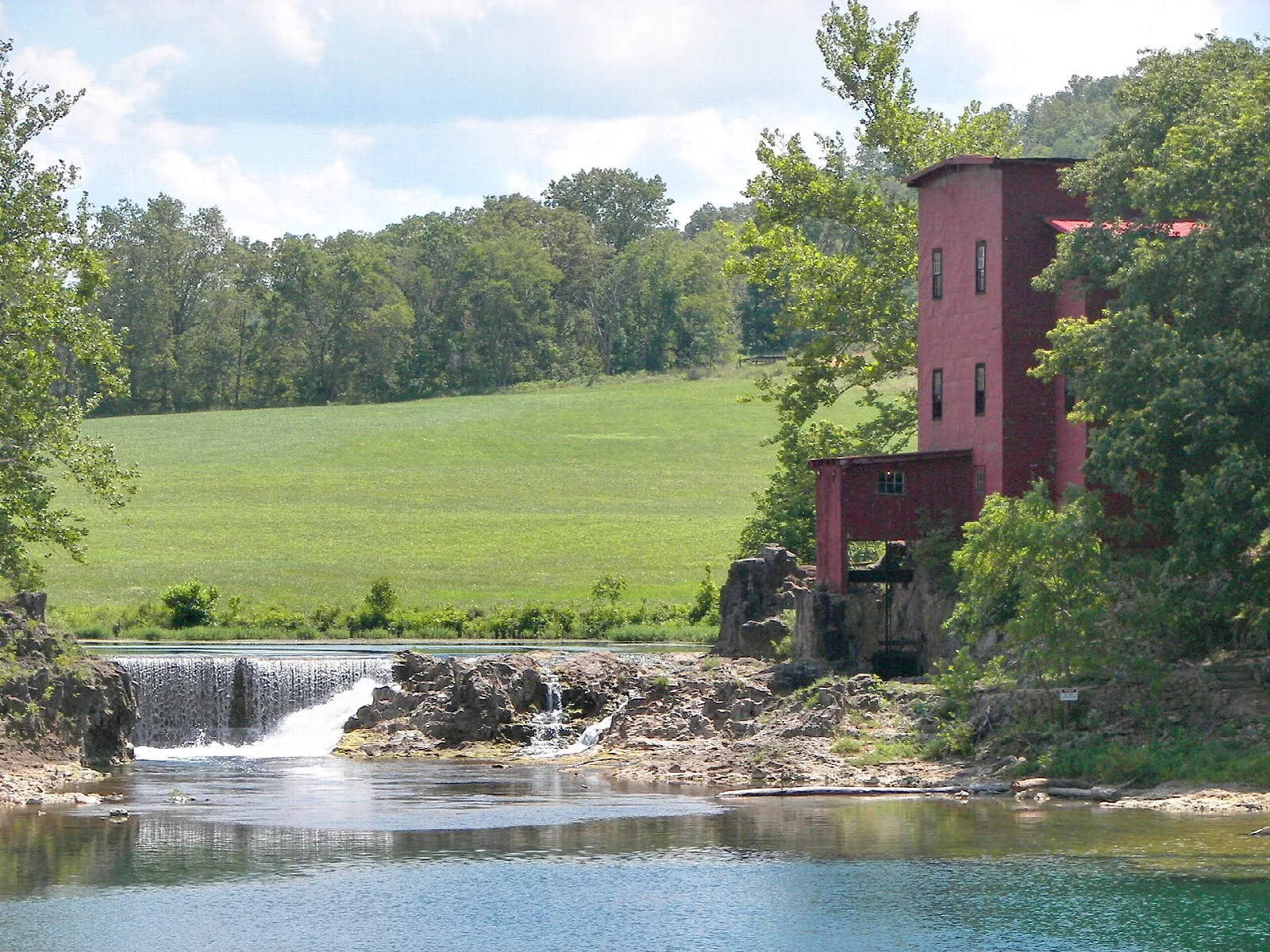 Red wooden mill beside a small waterfall and green pool, with fields behind