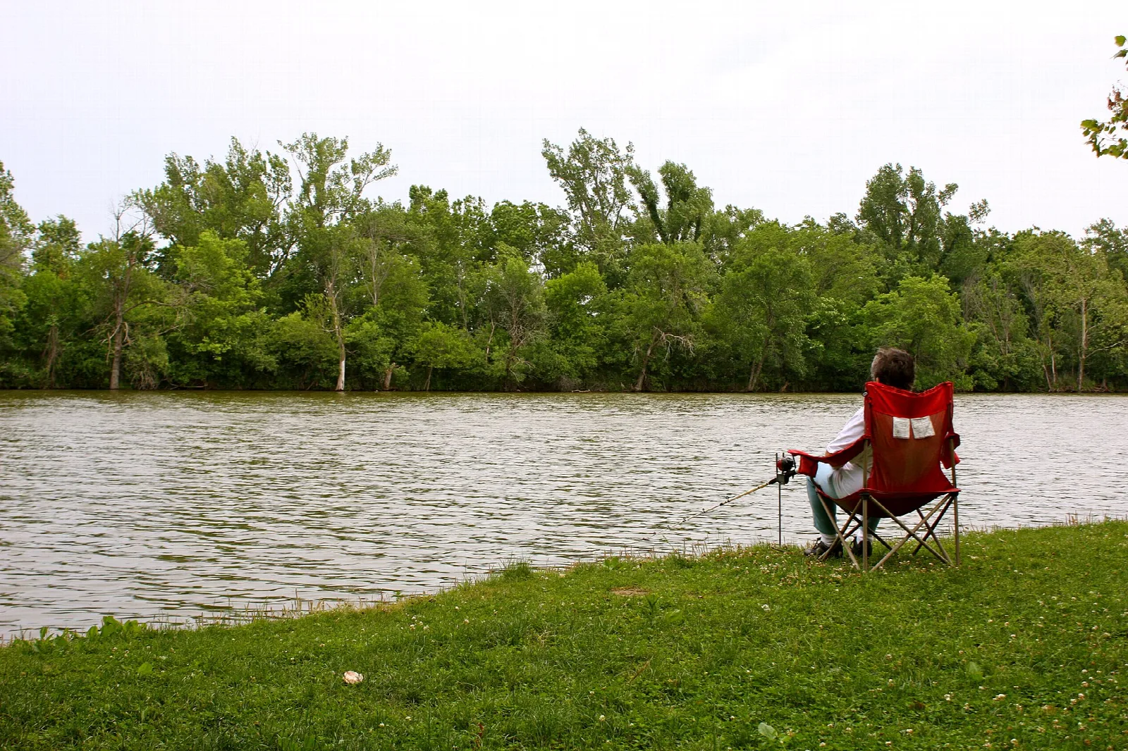 Person in a red chair fishing from a grassy lakeshore with trees across the water