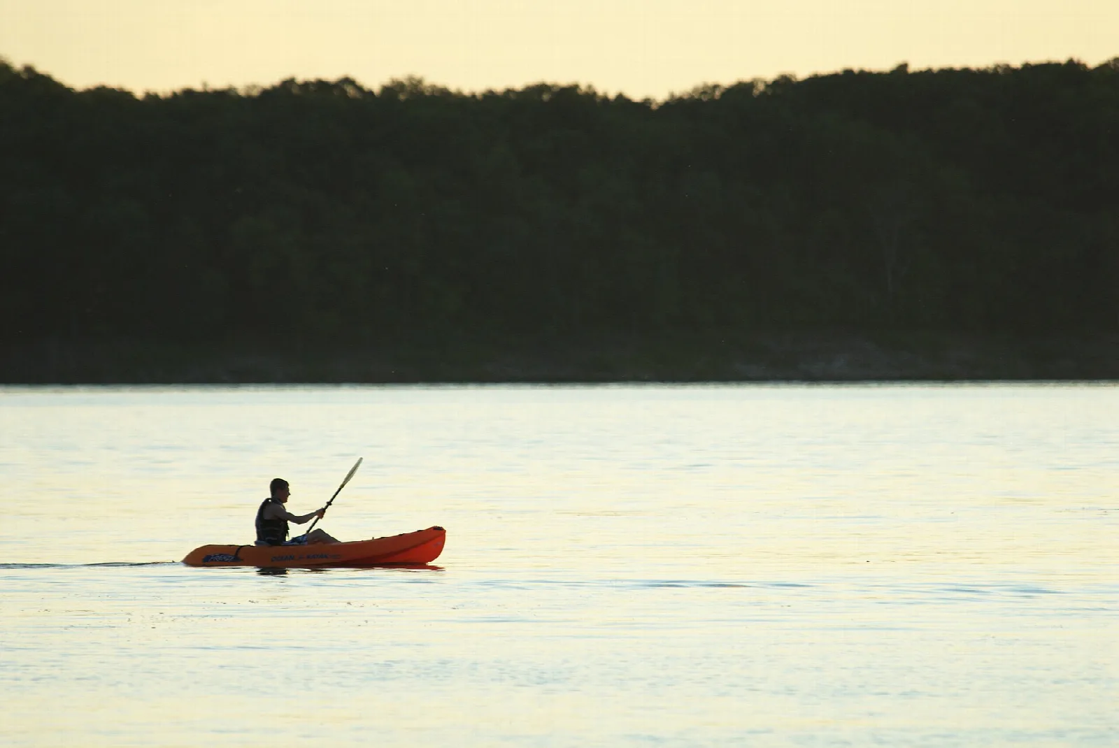 Person paddling a red kayak on calm water in front of a dark tree line