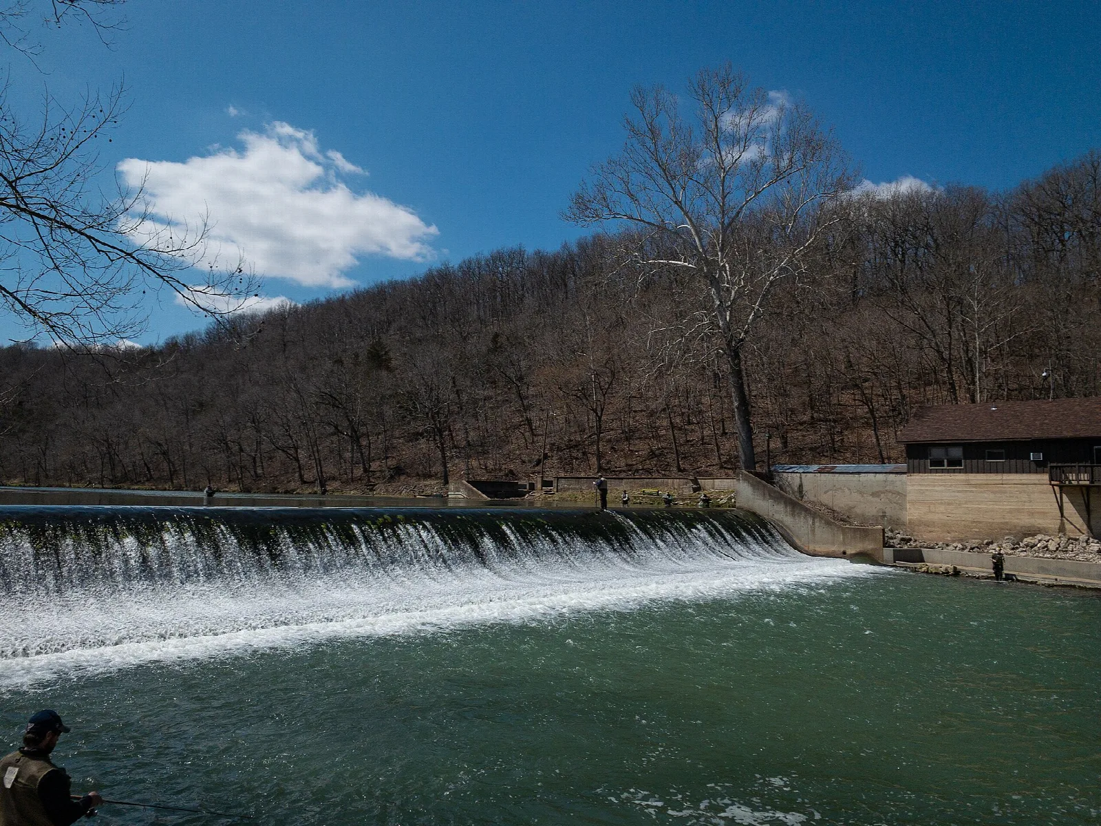 Low dam with water spilling over it, bare trees and a shelter beyond