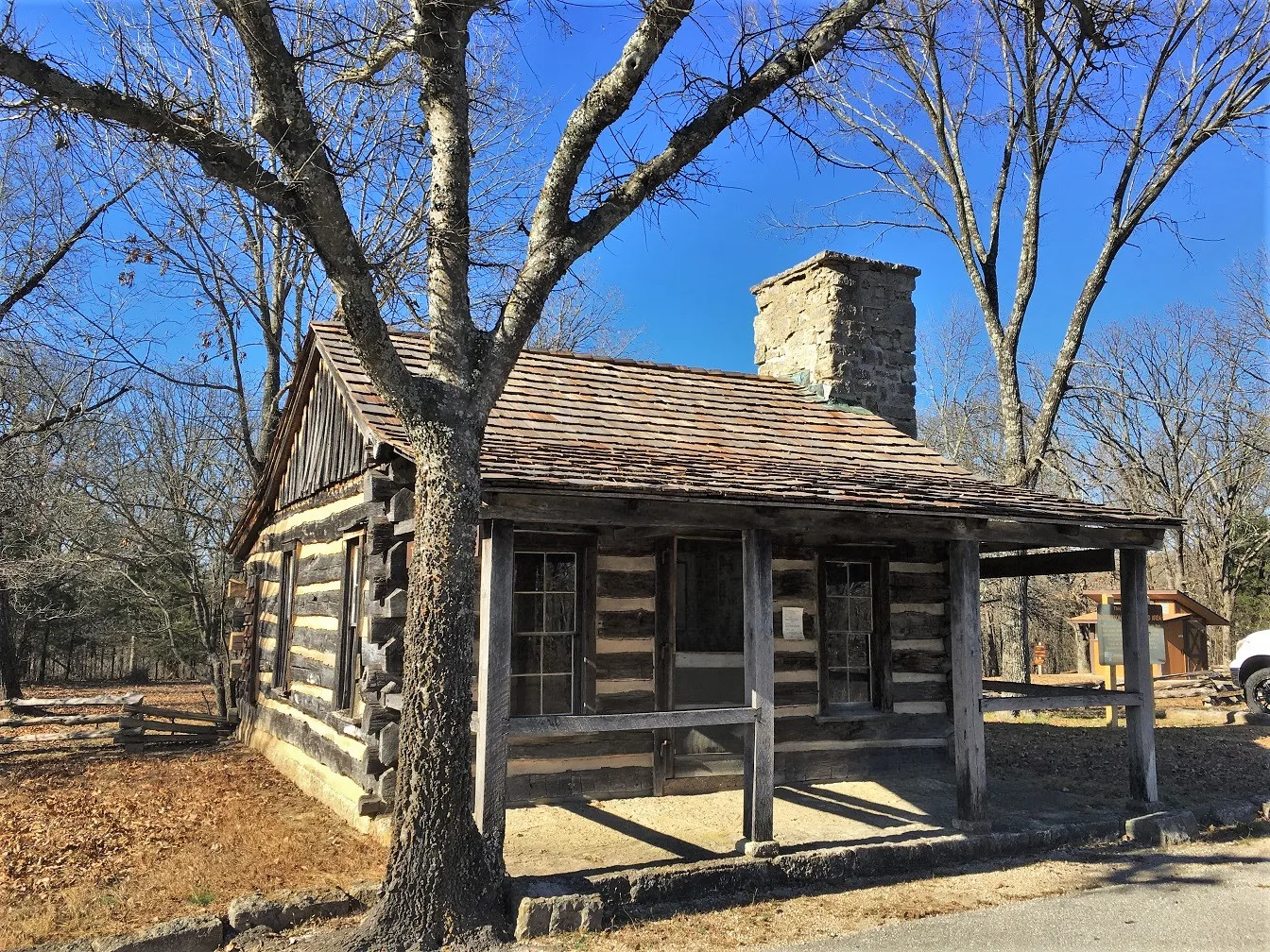Log cabin with a stone chimney and covered porch among bare trees