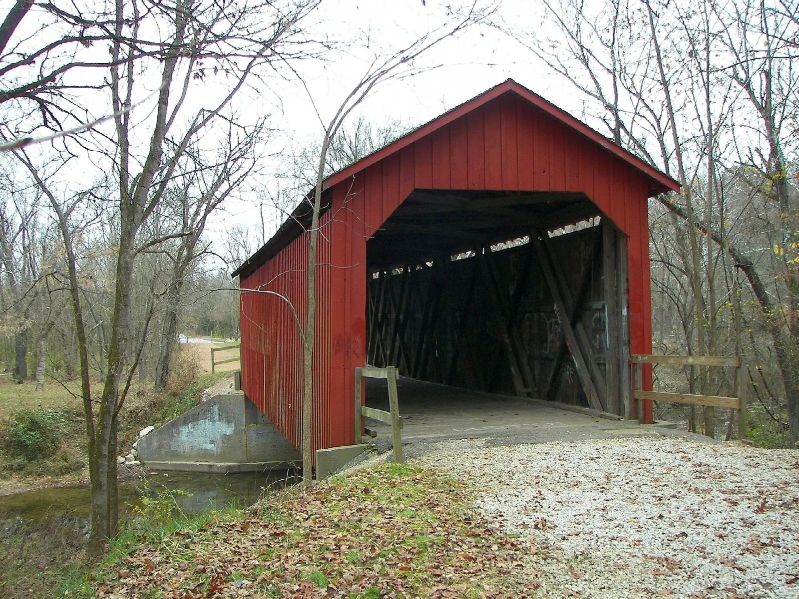 Red covered bridge over a creek with a gravel path and bare trees