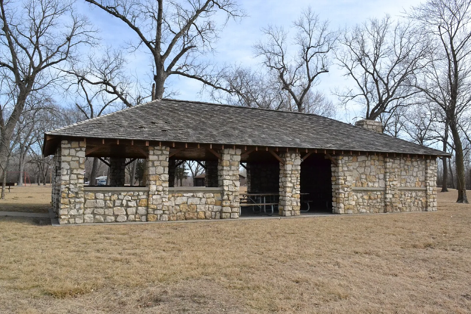 Open stone picnic shelter with a shingled roof on dry grass