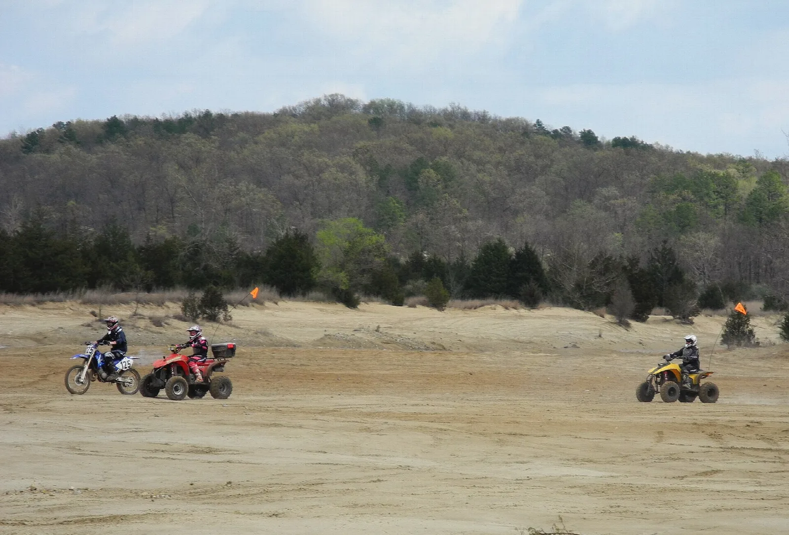 Riders on ATVs and a motorcycle crossing open sand with wooded hills behind