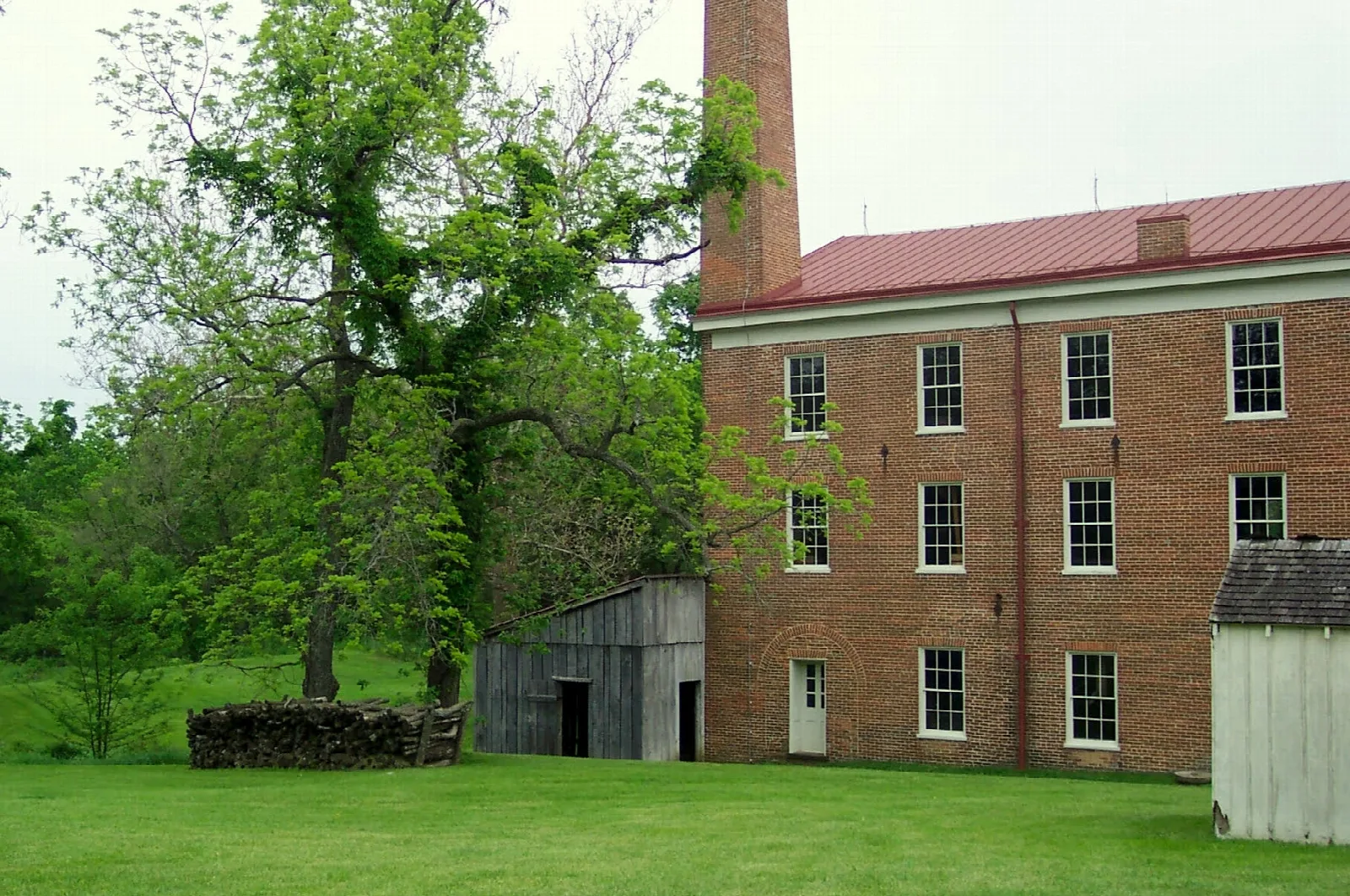Multi-story brick mill building and chimney beside green lawn and trees