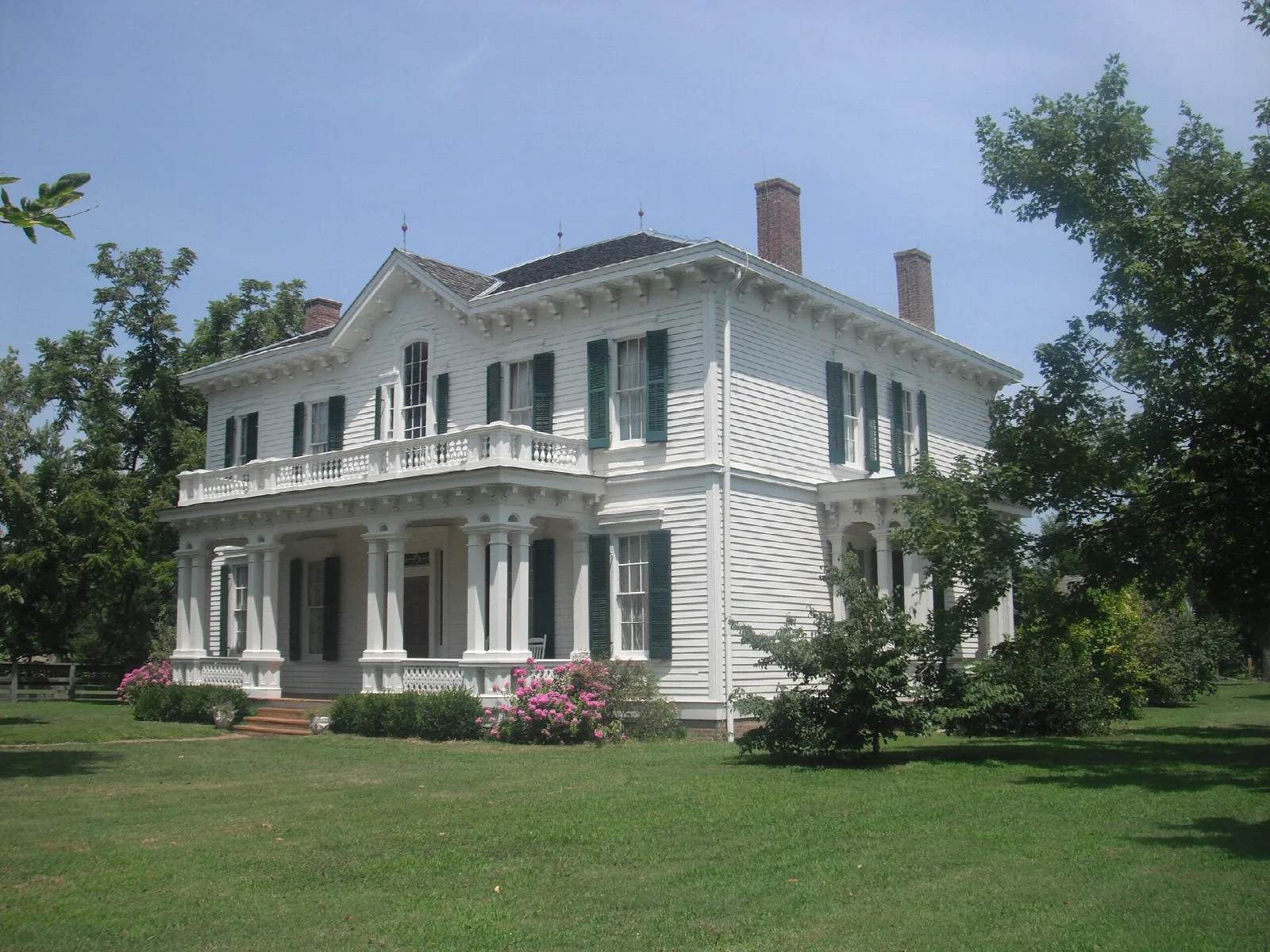 White two-story historic house with balconies and green shutters behind a lawn