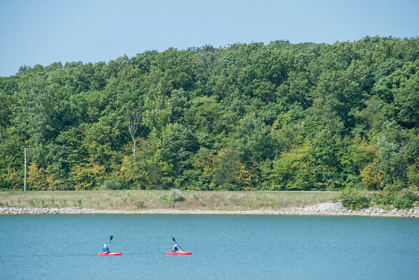 Two people in red kayaks on blue water below a wooded hillside