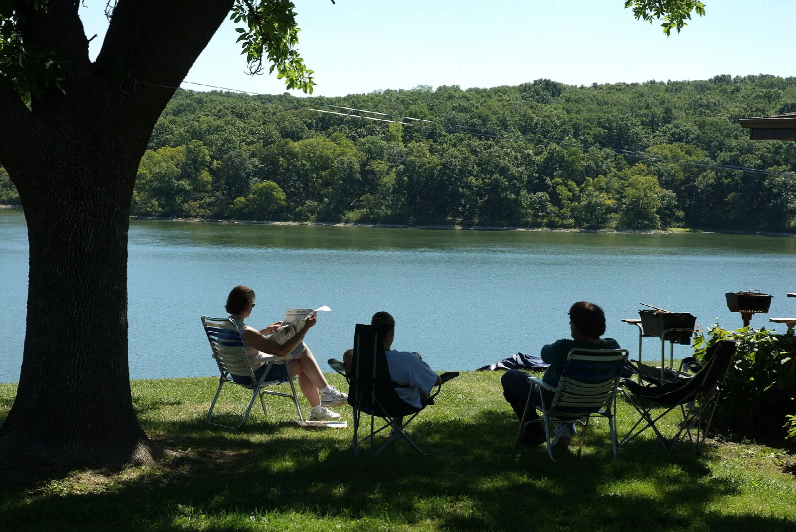 Three people in lawn chairs under a tree looking over a wooded lake