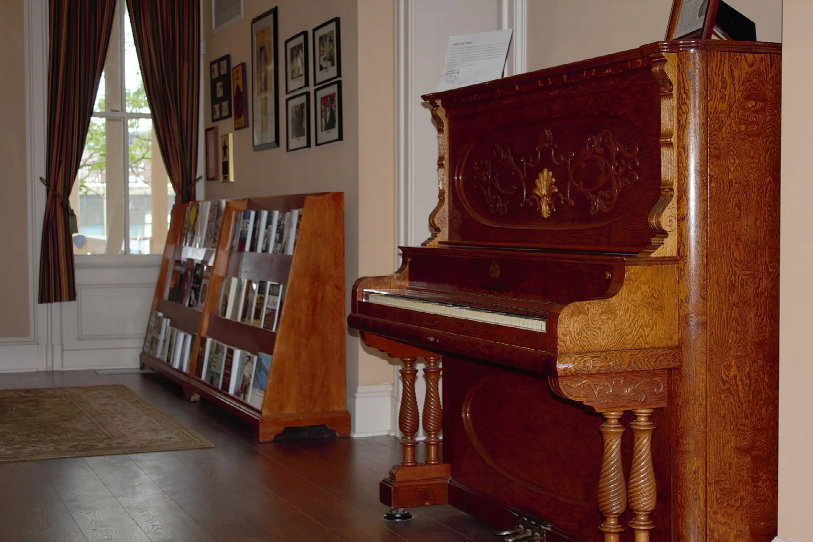 Upright wooden piano beside a bookshelf and curtained window