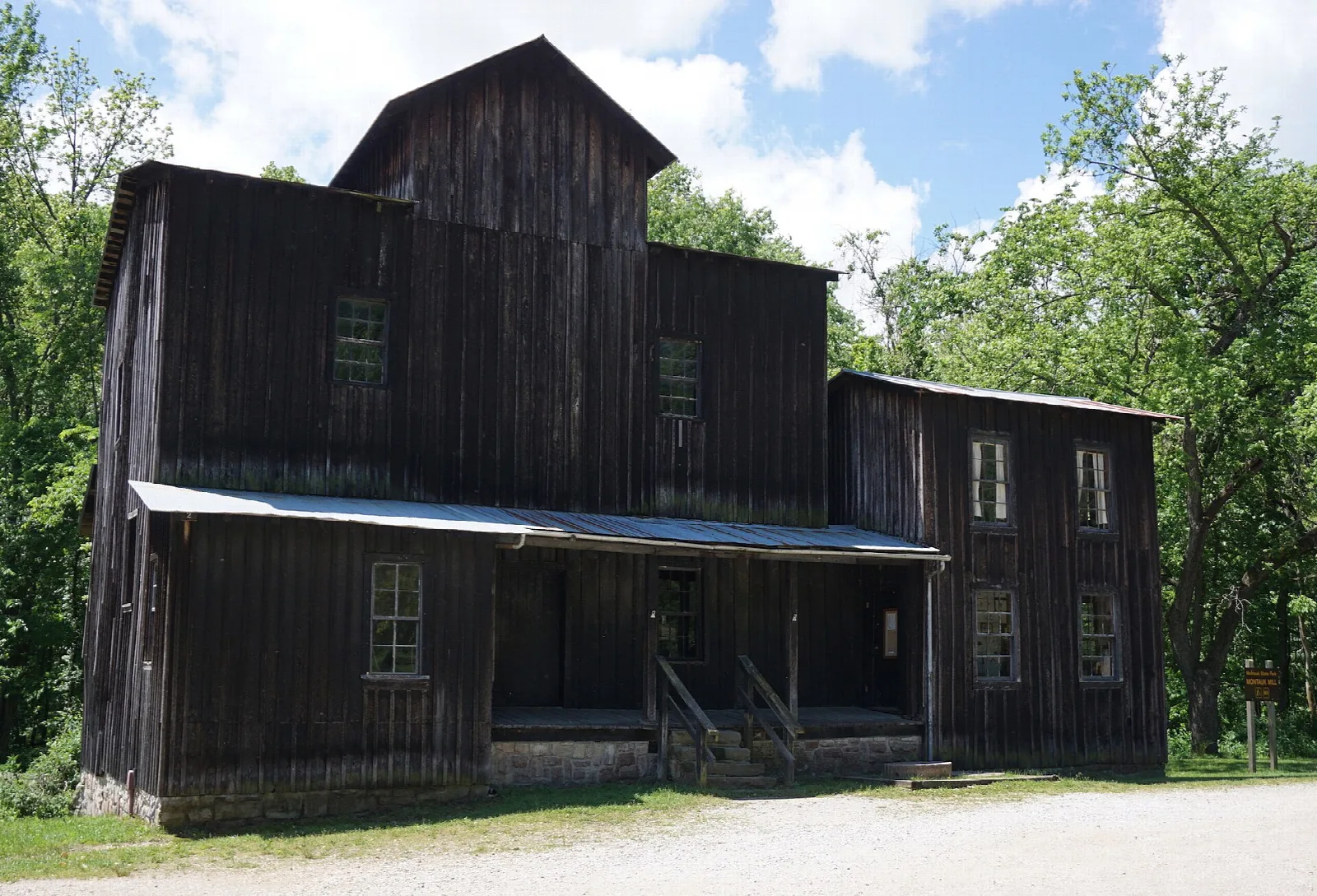Tall dark wooden mill building beside a gravel lot and green trees