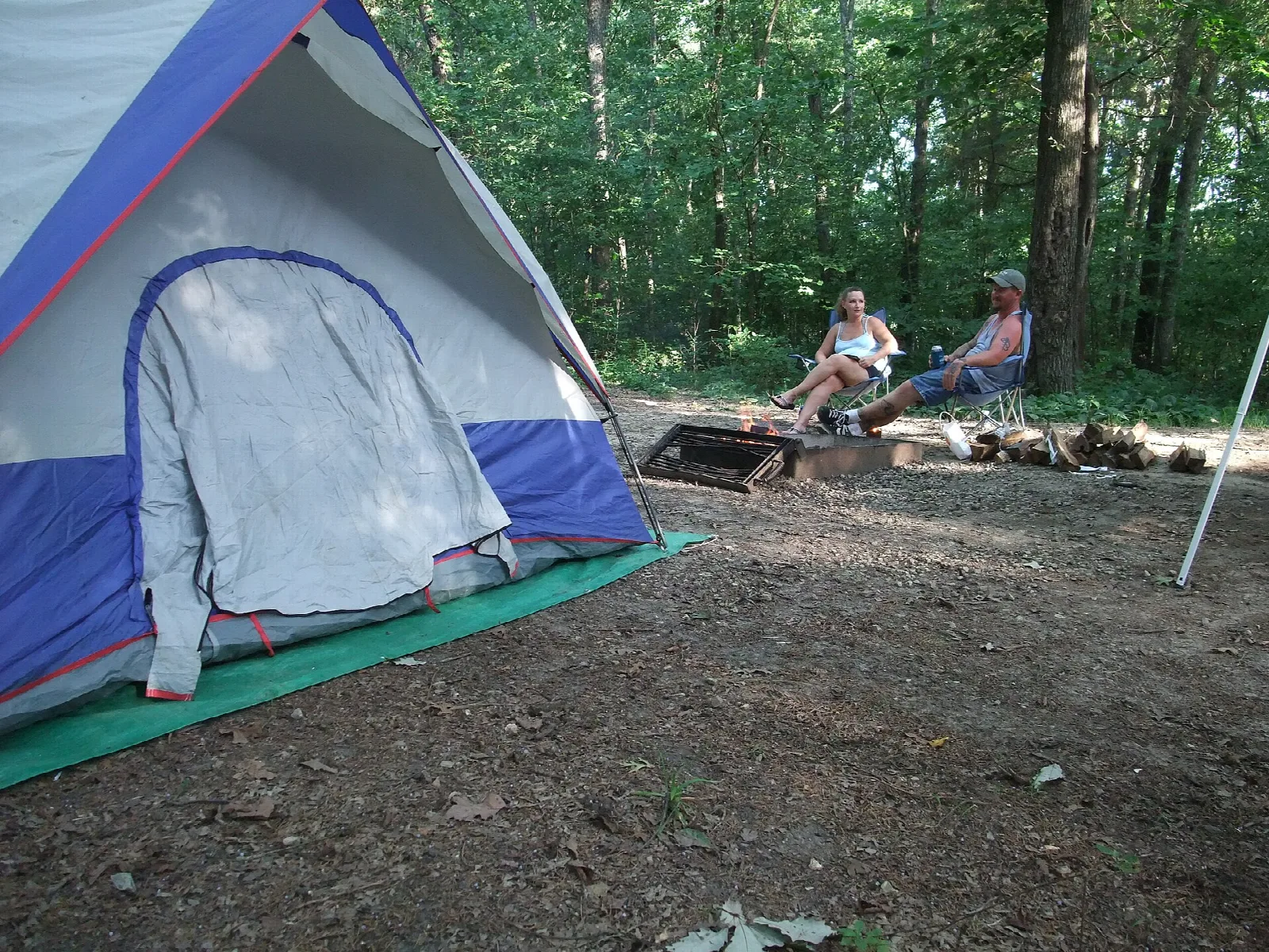 Blue and white tent at a wooded campsite with two people in chairs