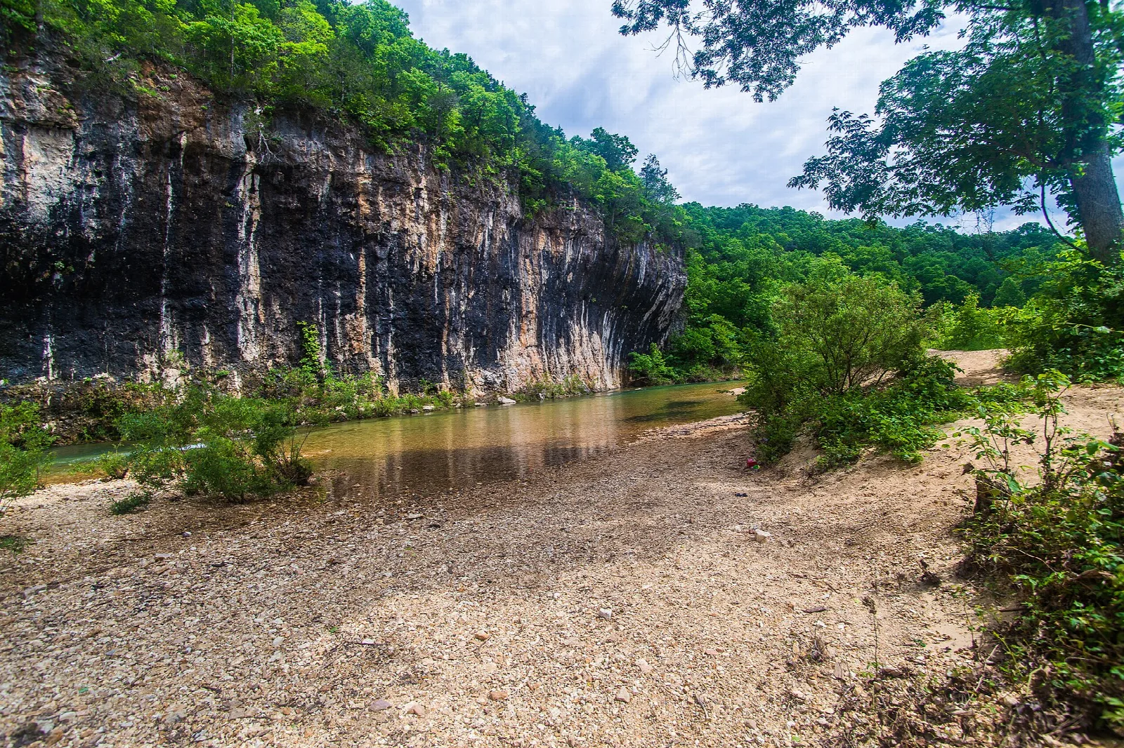 Tall rock bluff beside a shallow creek and gravel bar, with green trees above