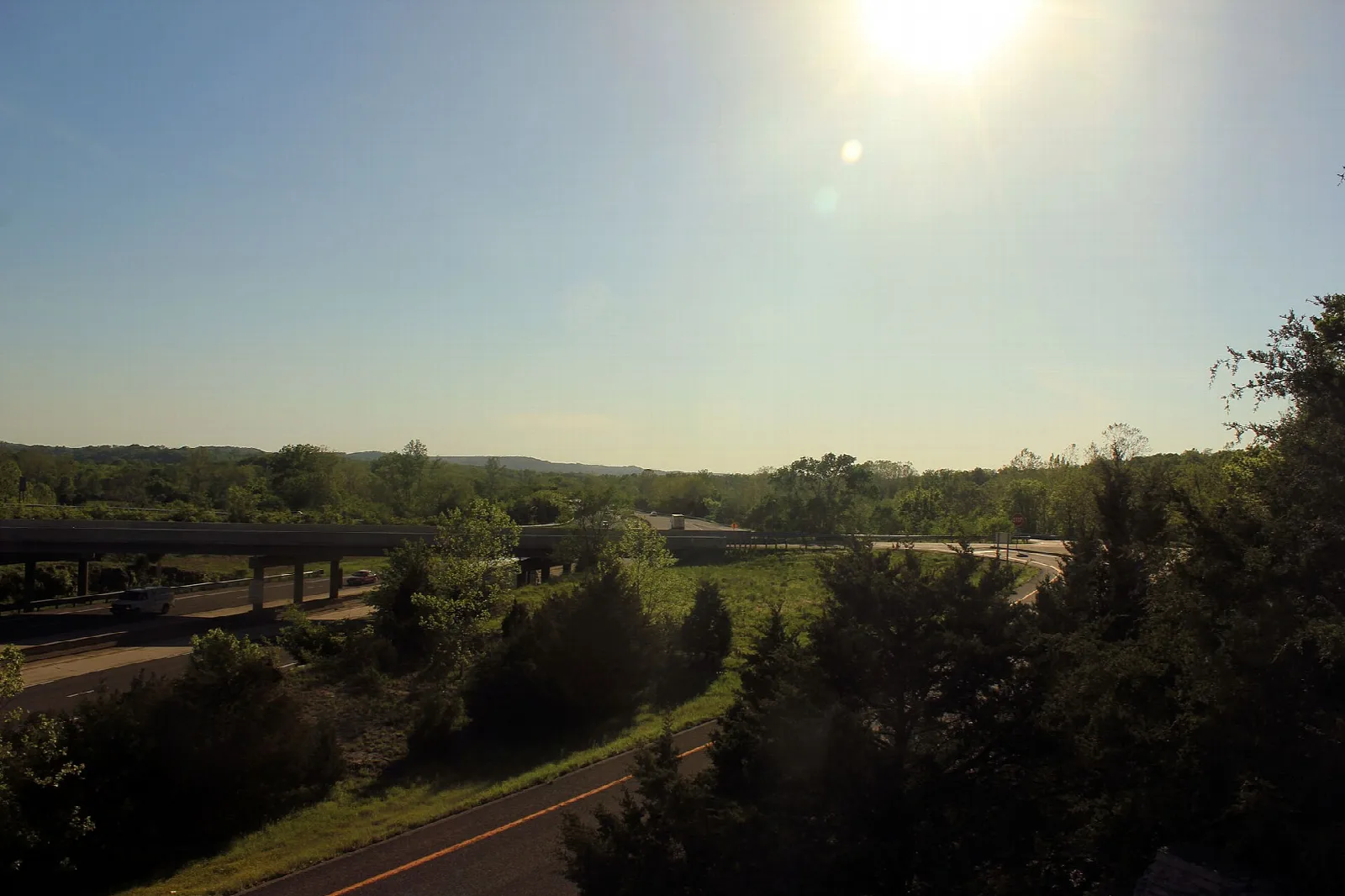 Low sun over green wooded hills with a roadway and bridge in the distance