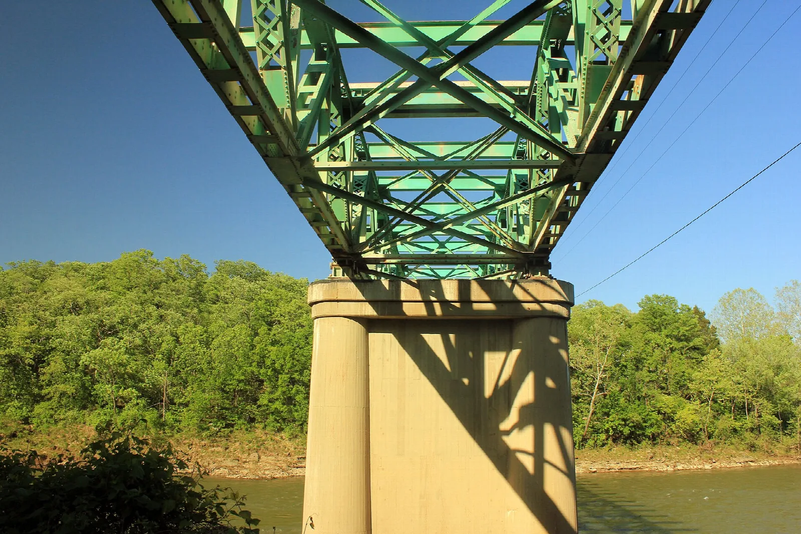 Green steel bridge truss and concrete pier above a river bank with trees