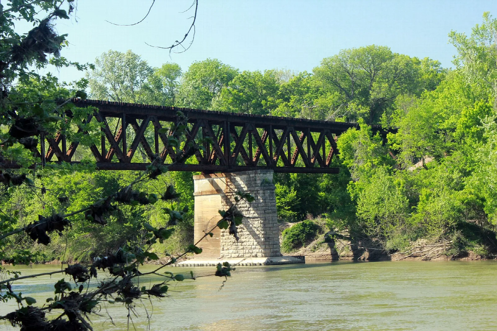 Old steel truss bridge and stone pier over a wide river framed by green trees
