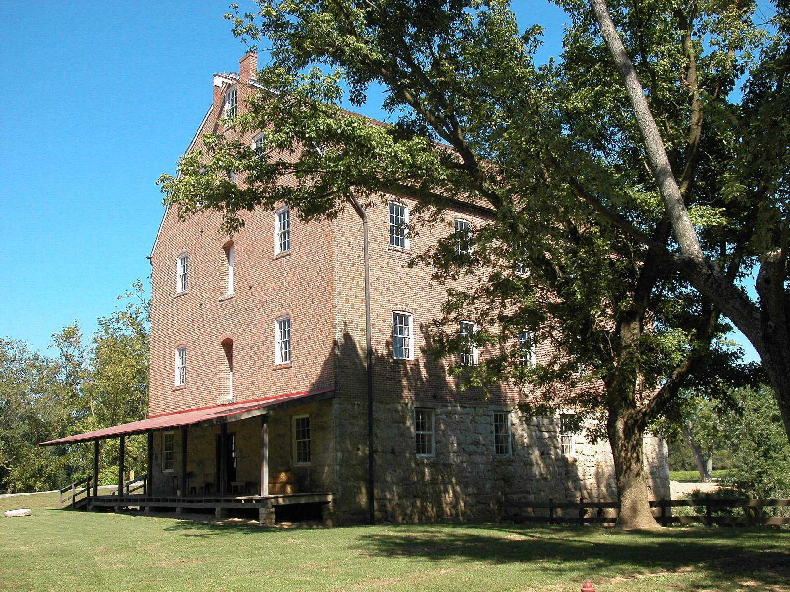 Tall brick mill with a red roof porch under a large tree