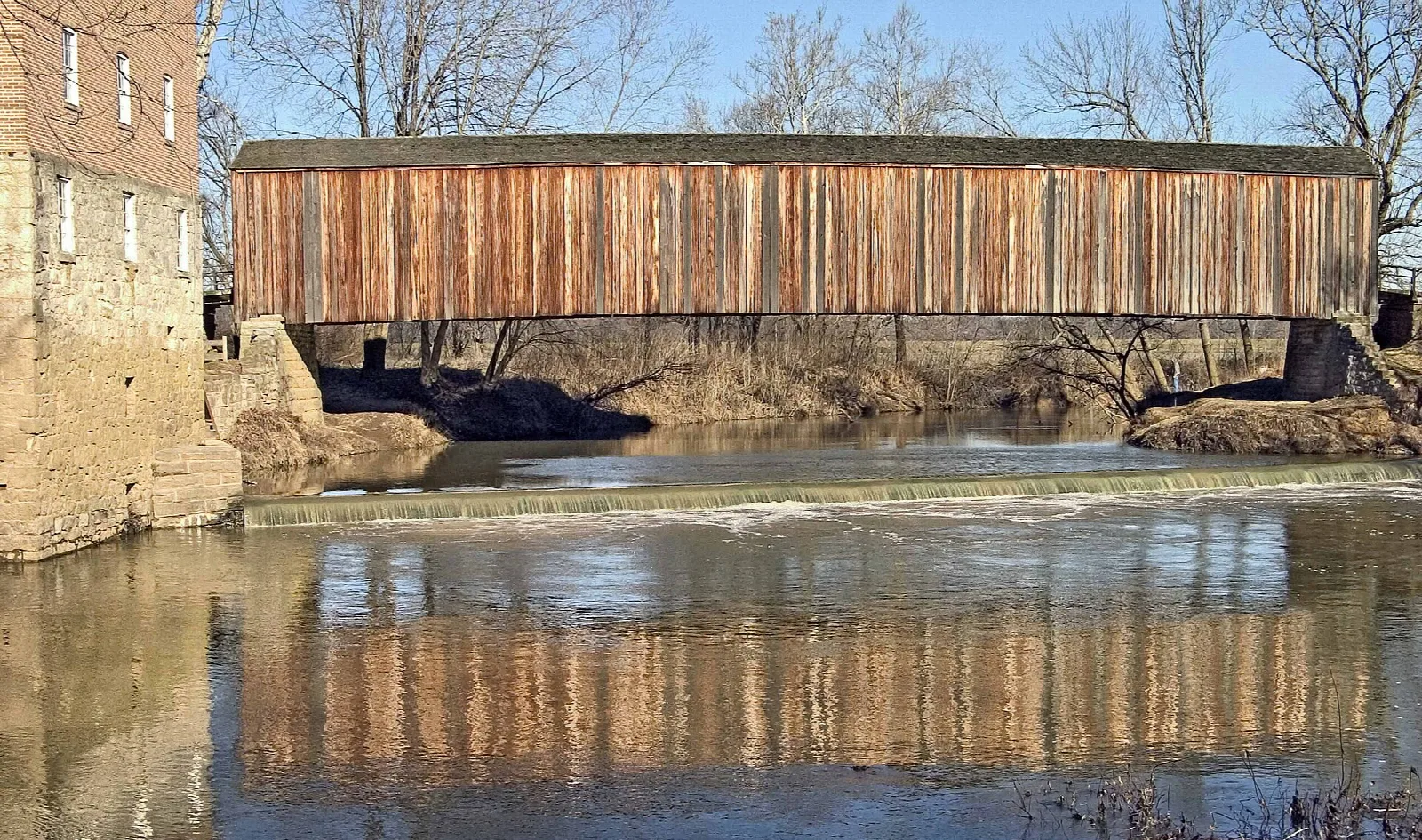 Wooden covered bridge and stone mill wall reflected in a river