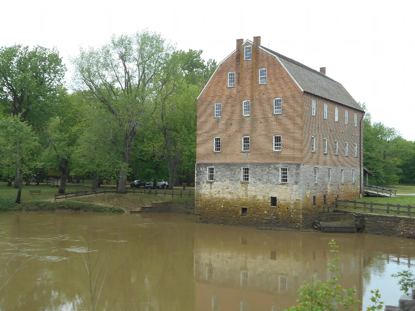 Tall brick and stone mill beside a brown river with trees behind