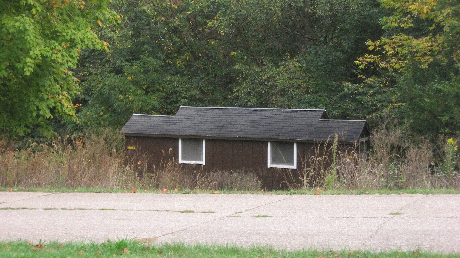 Small dark wooden building with white windows beside a paved lot, in front of trees